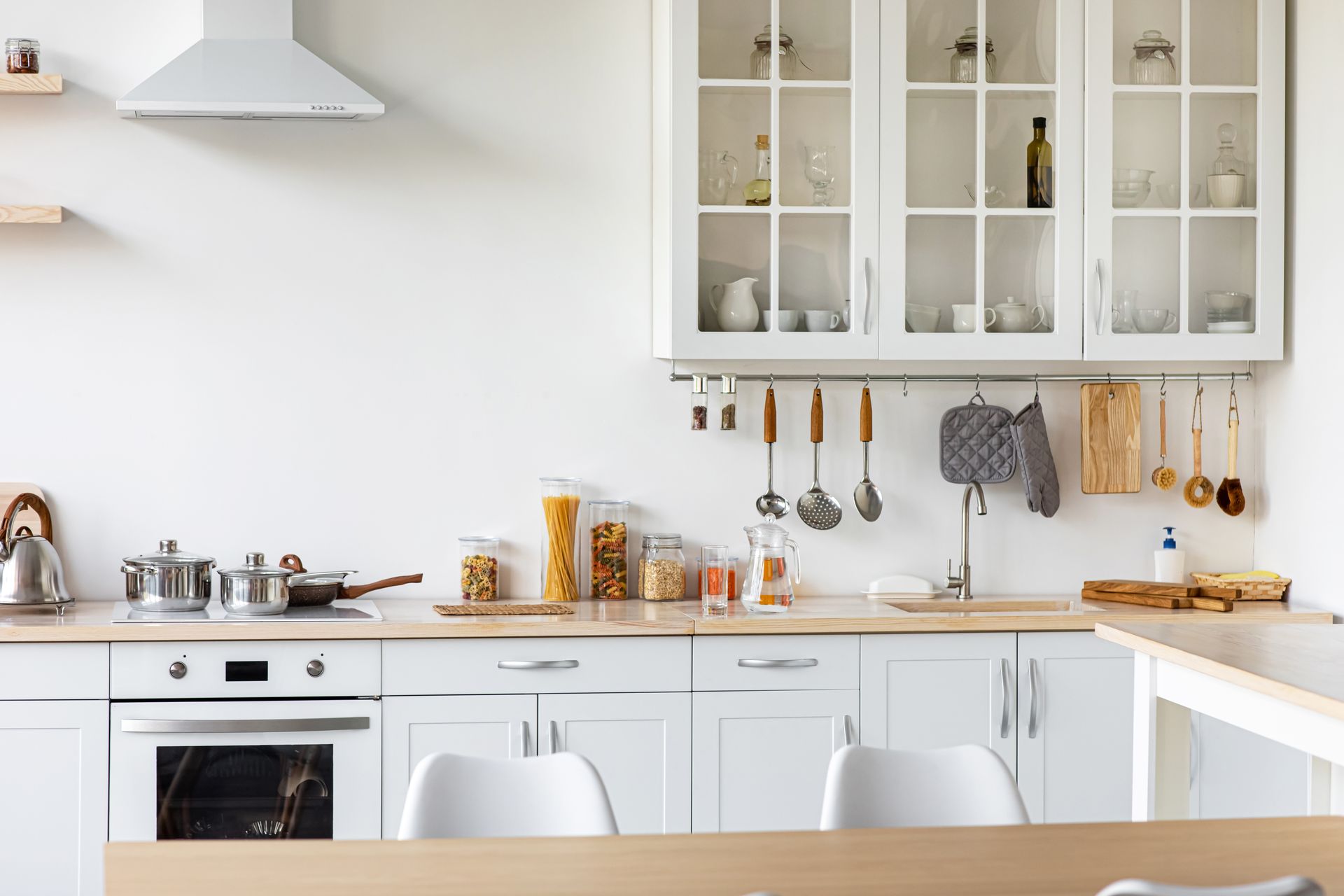 Bright white kitchen with cabinets, oven, hanging utensils, and a countertop with jars and cookware