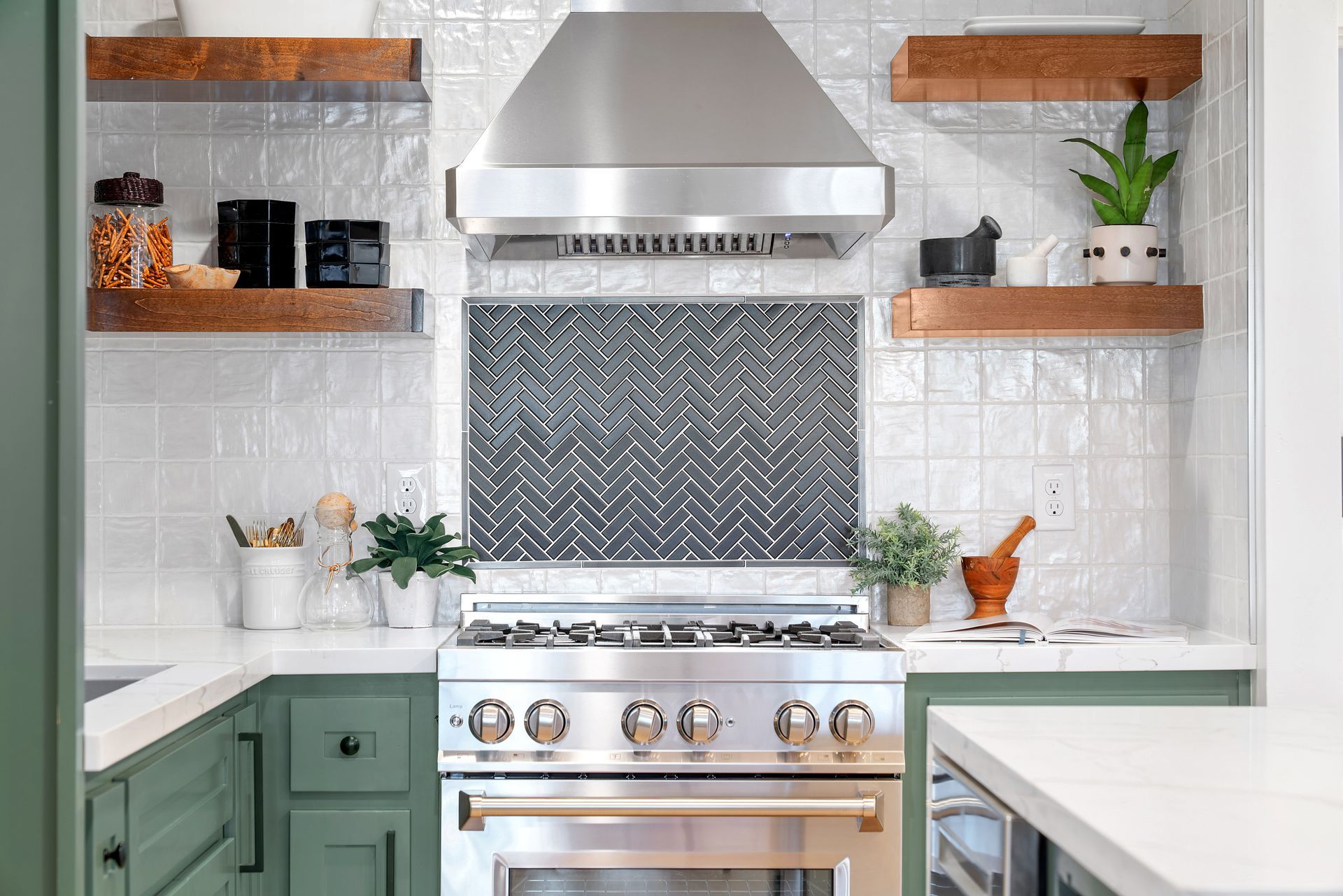 Modern white kitchen with green cabinets, stainless range hood, herringbone backsplash, and open shelves