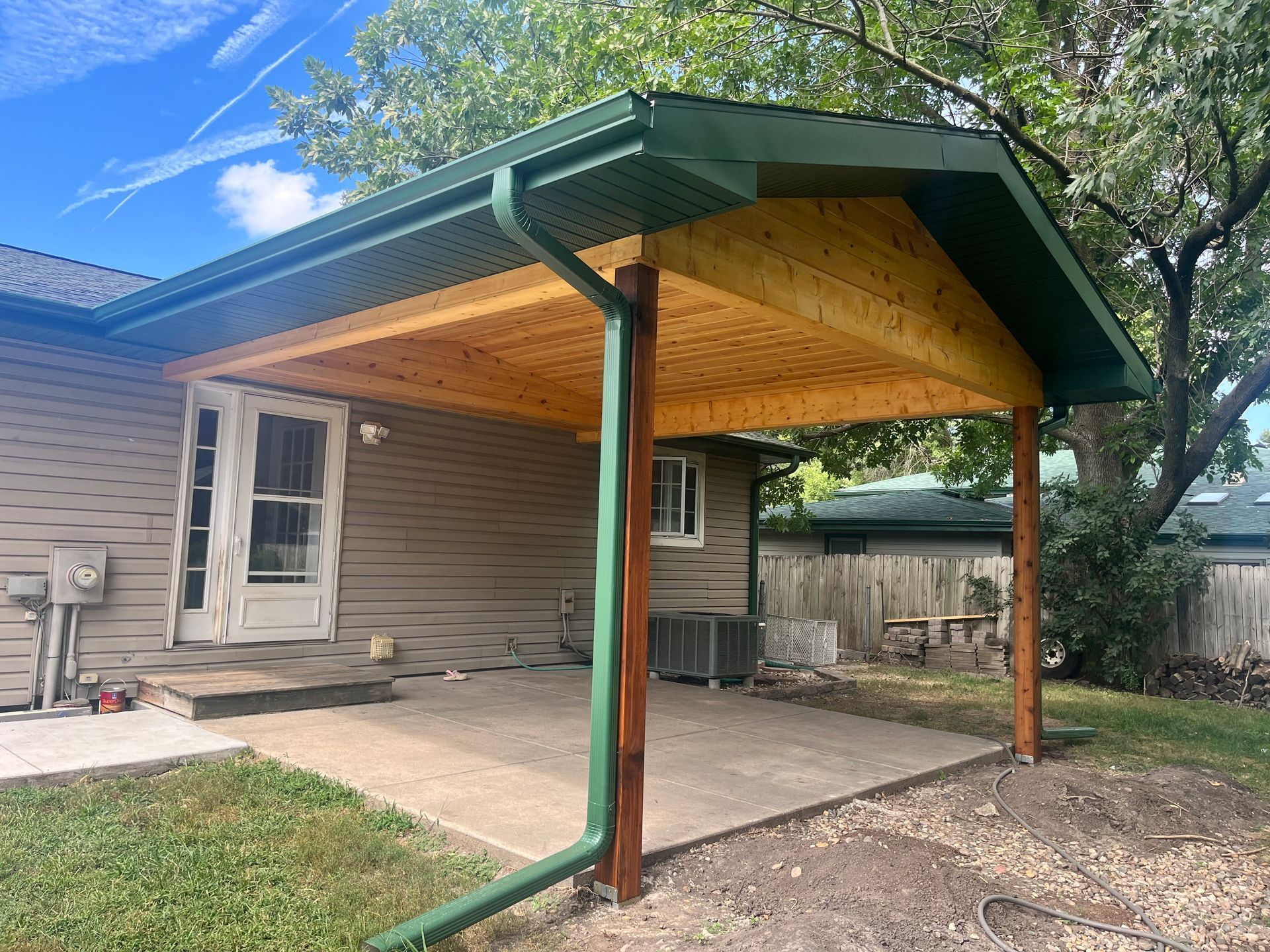 Covered patio with green roof and wood ceiling attached to a house, overlooking a concrete slab and yard
