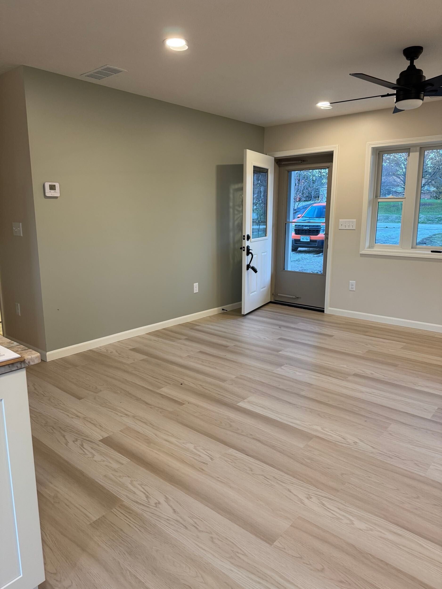 Empty living room with light wood floors, beige walls, ceiling fan, and open front door.
