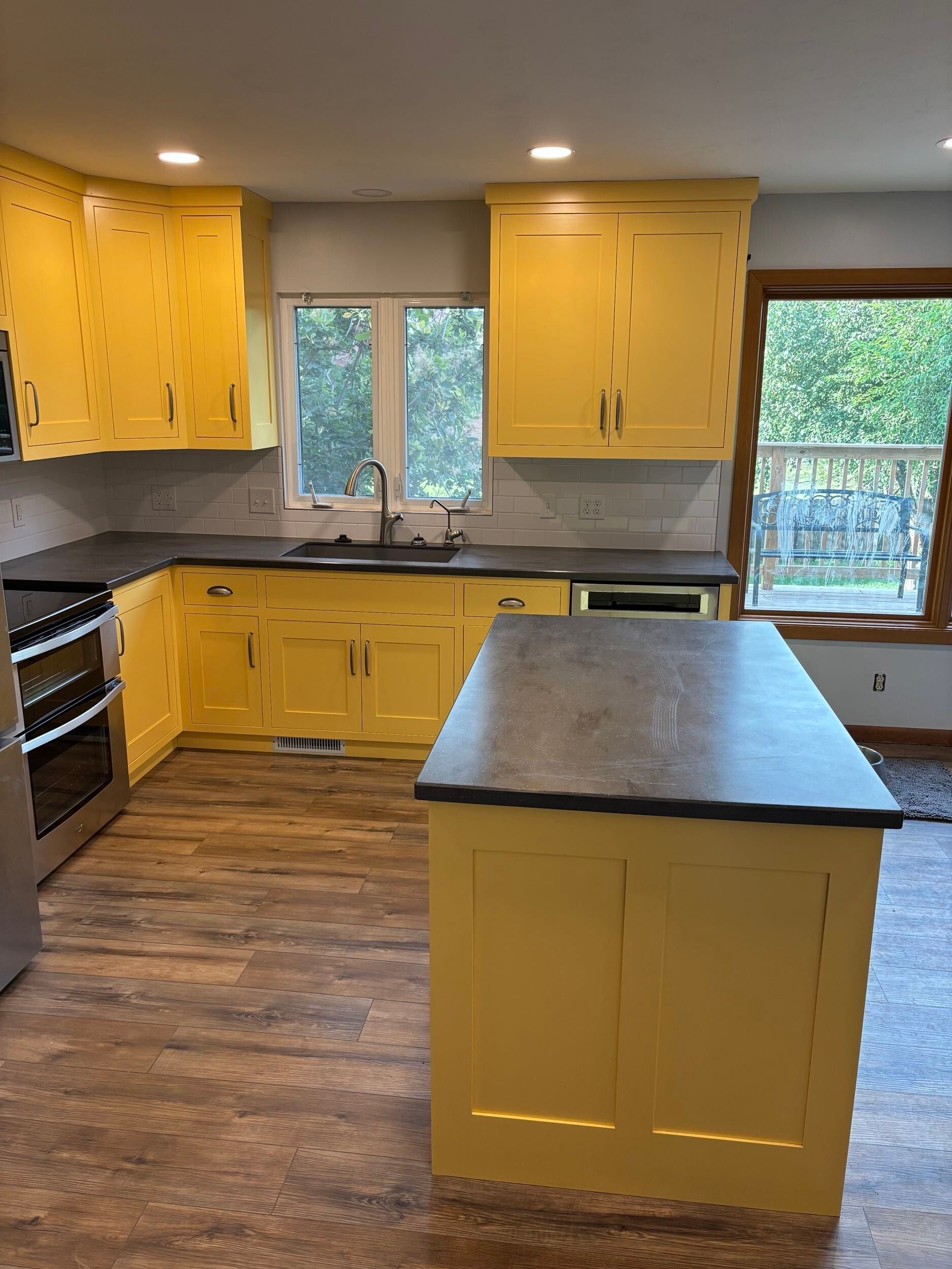 Bright yellow kitchen with black countertops, central island, and wood flooring in front of large windows