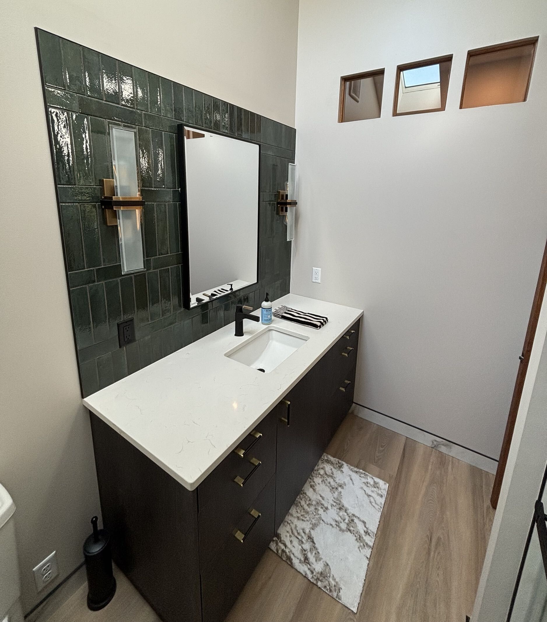Modern bathroom with dark vanity, large mirror, white sink, and three small frosted windows.