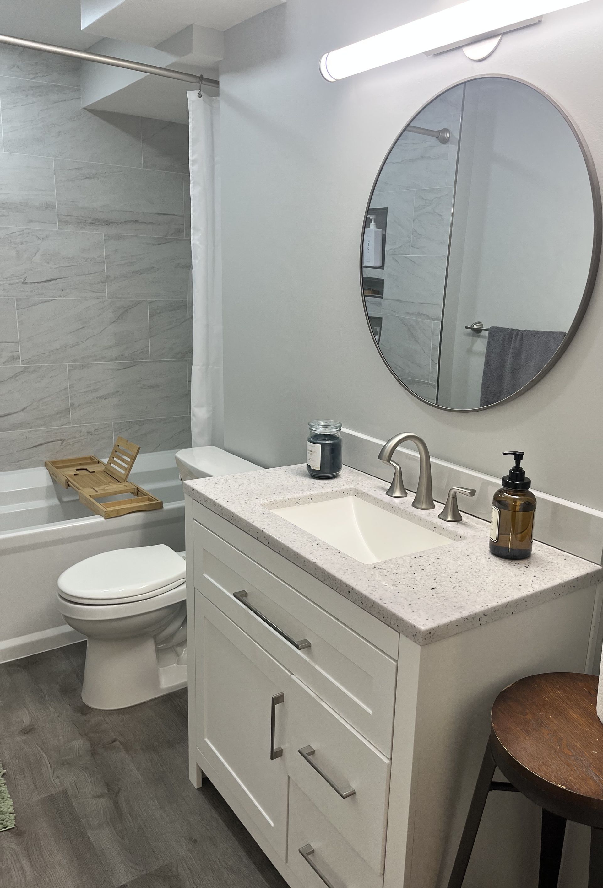 Bright modern bathroom with white vanity, round mirror, toilet, and tiled shower.