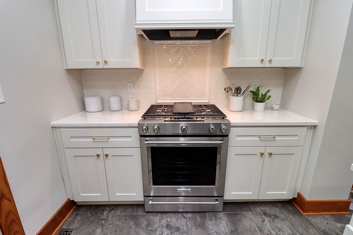 White kitchen with stainless steel stove, range hood, and cabinets around a gray tiled backsplash and floor