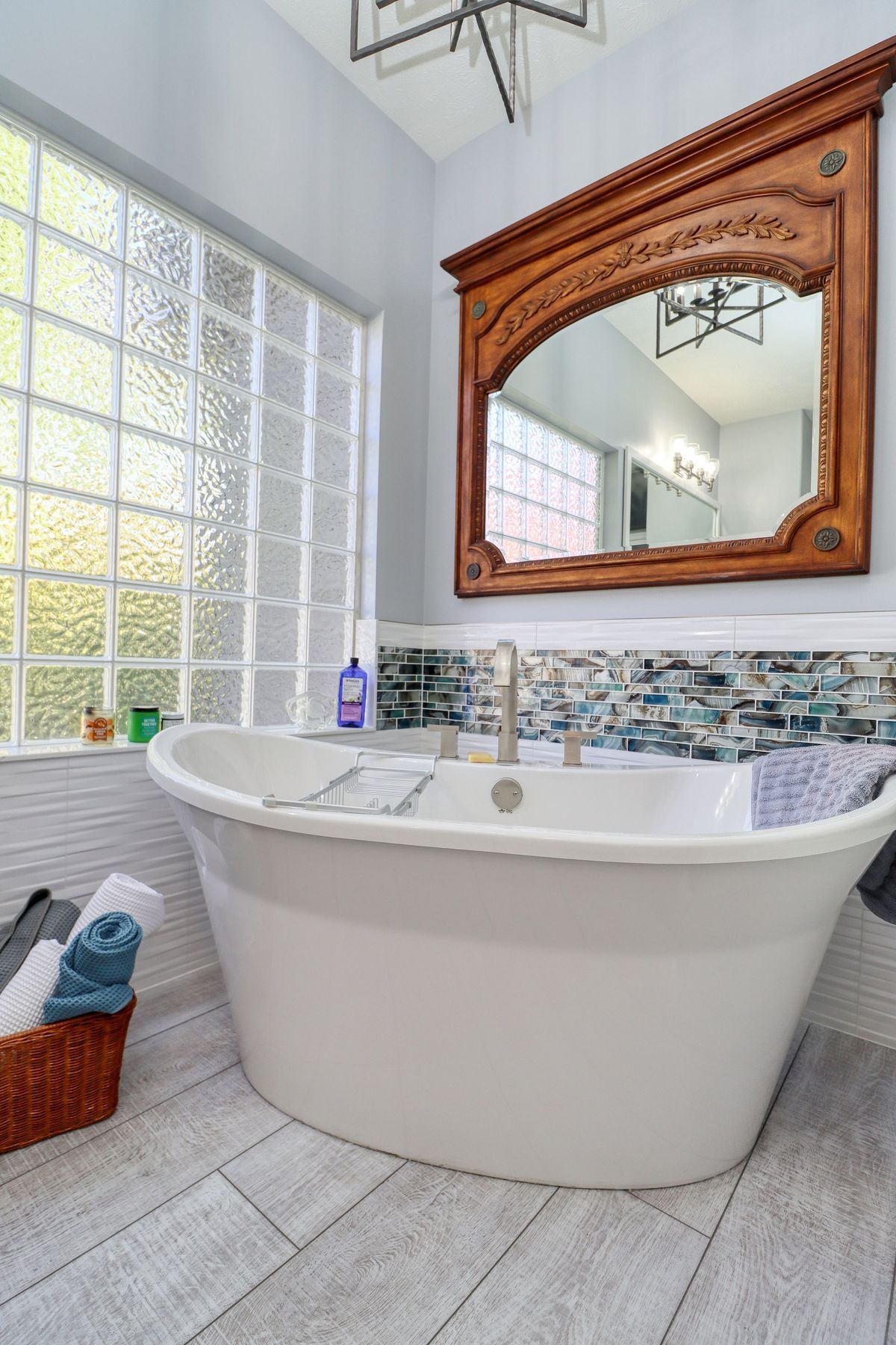 Modern bathroom with a white soaking tub, wooden mirror, glass block window, and gray tile floor