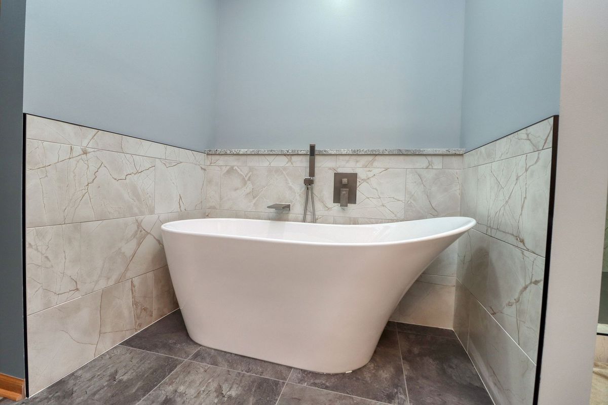 Modern freestanding bathtub in a tiled bathroom nook with light blue walls and gray floor tiles