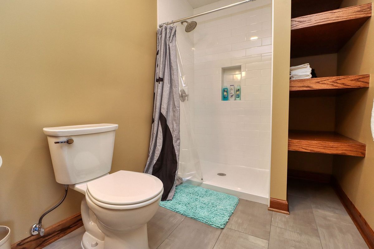 Small bathroom with toilet, shower, turquoise bath mat, and open shelving with baskets.