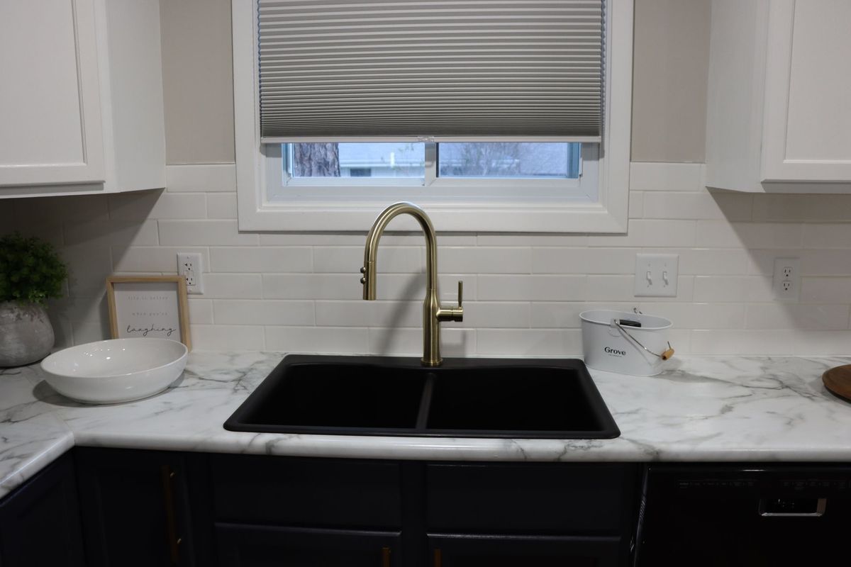 Modern kitchen sink with brass faucet, white countertops, and a window with a closed blind.