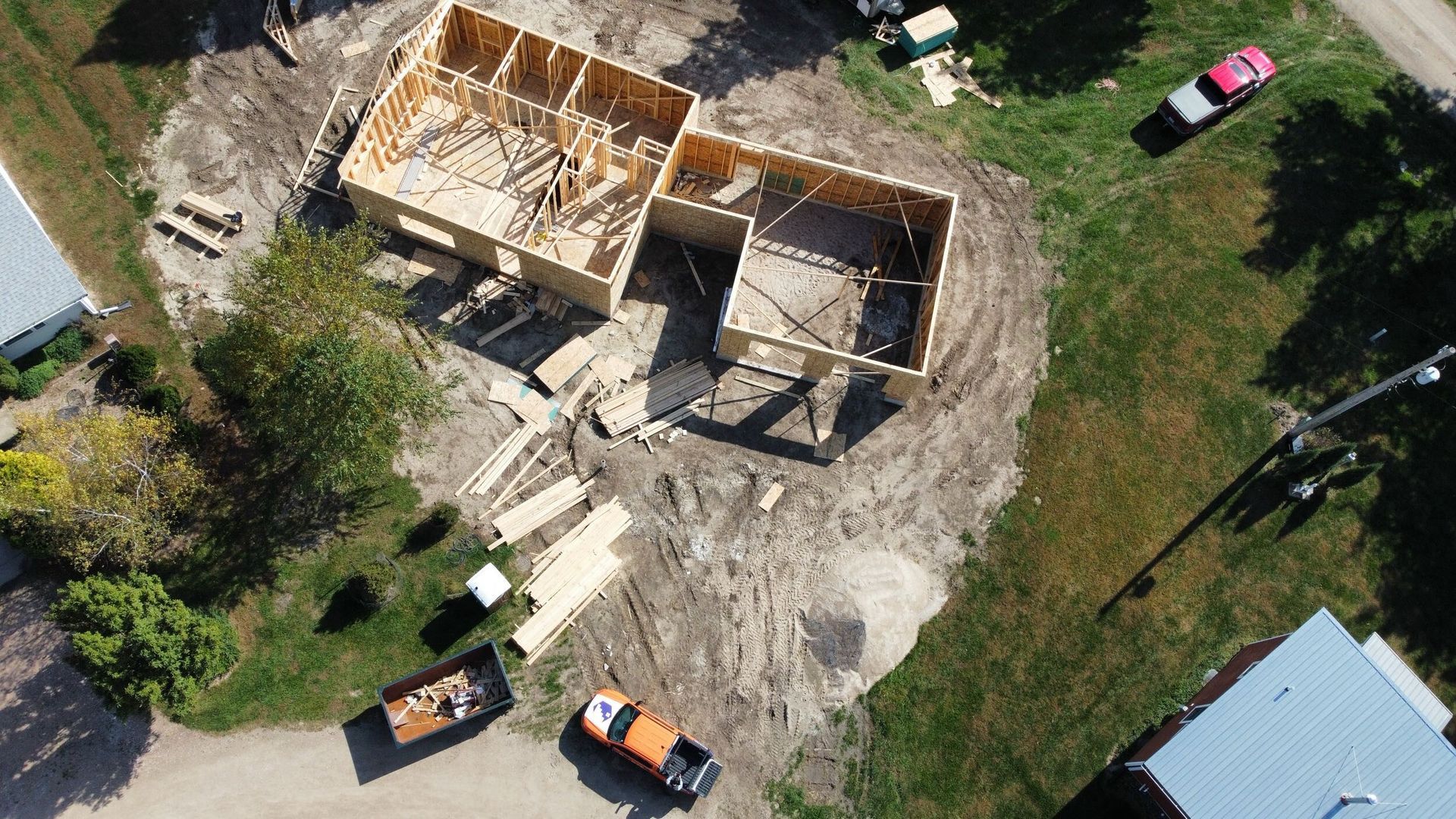Aerial view of a house under construction with lumber framing, trucks, and building materials on dirt lot.