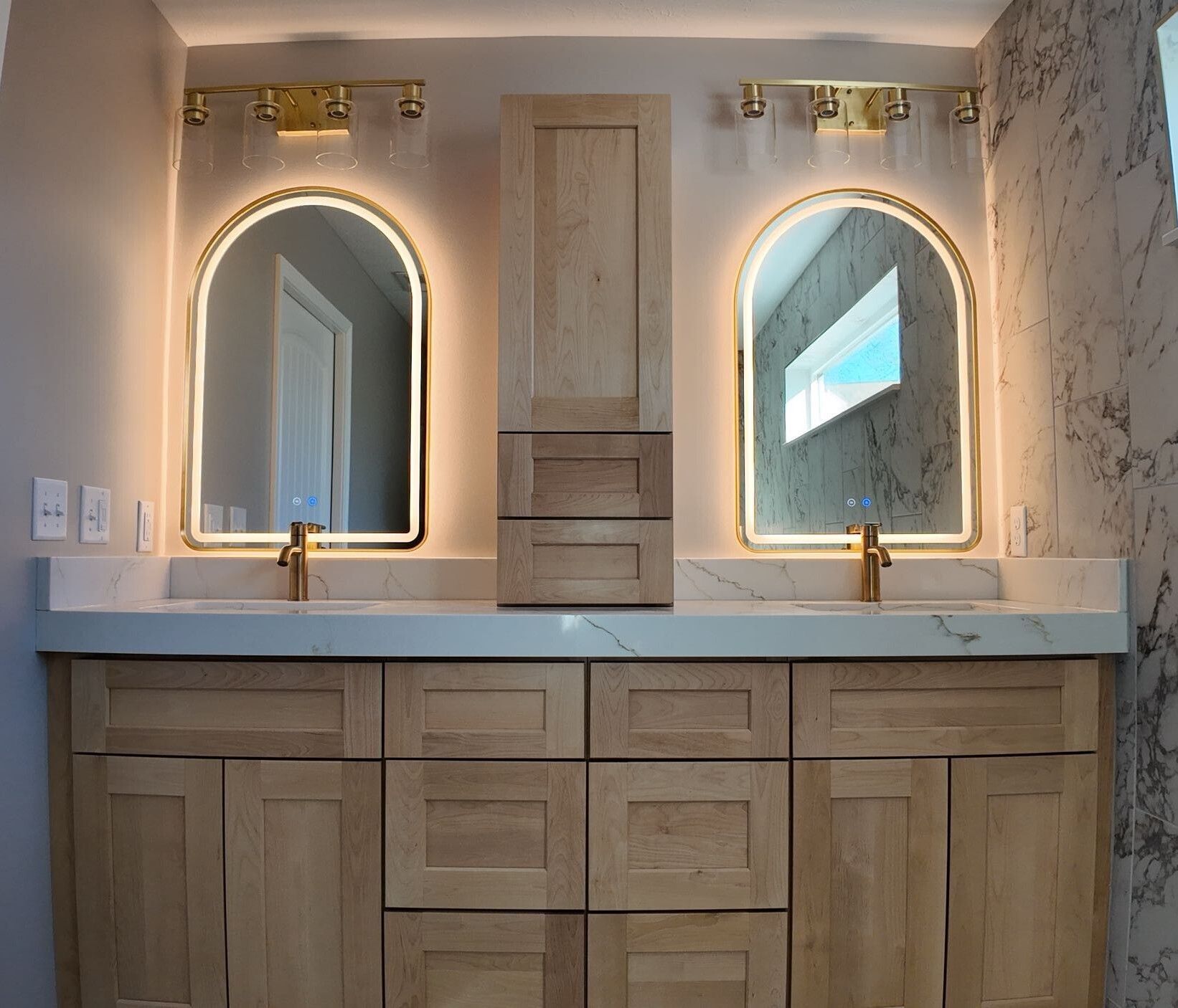 Double vanity bathroom with lit mirrors, white countertop, wood cabinets, and marble tile walls