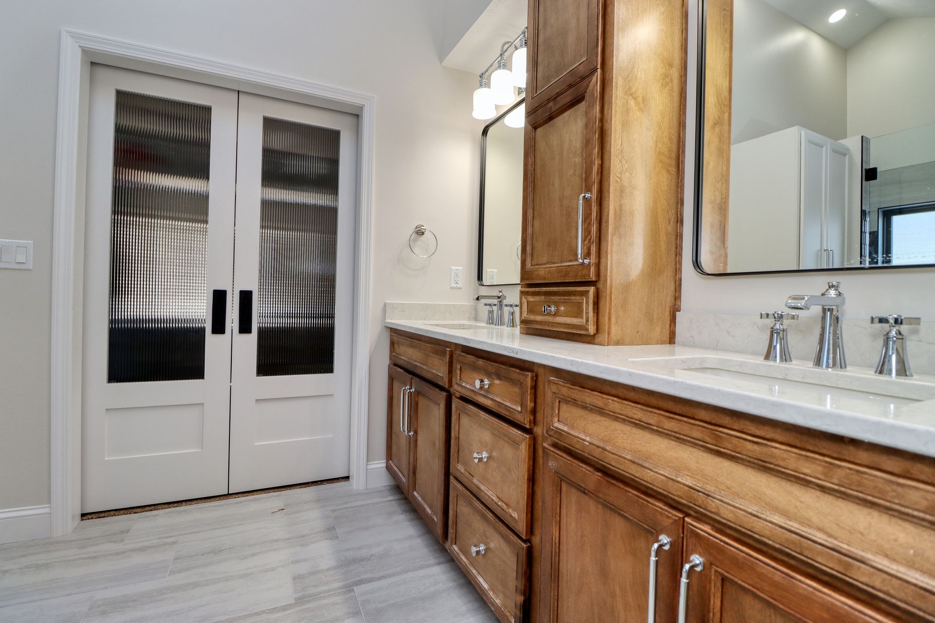 Bright bathroom with double vanity, wood cabinets, white counters, and French doors with frosted glass