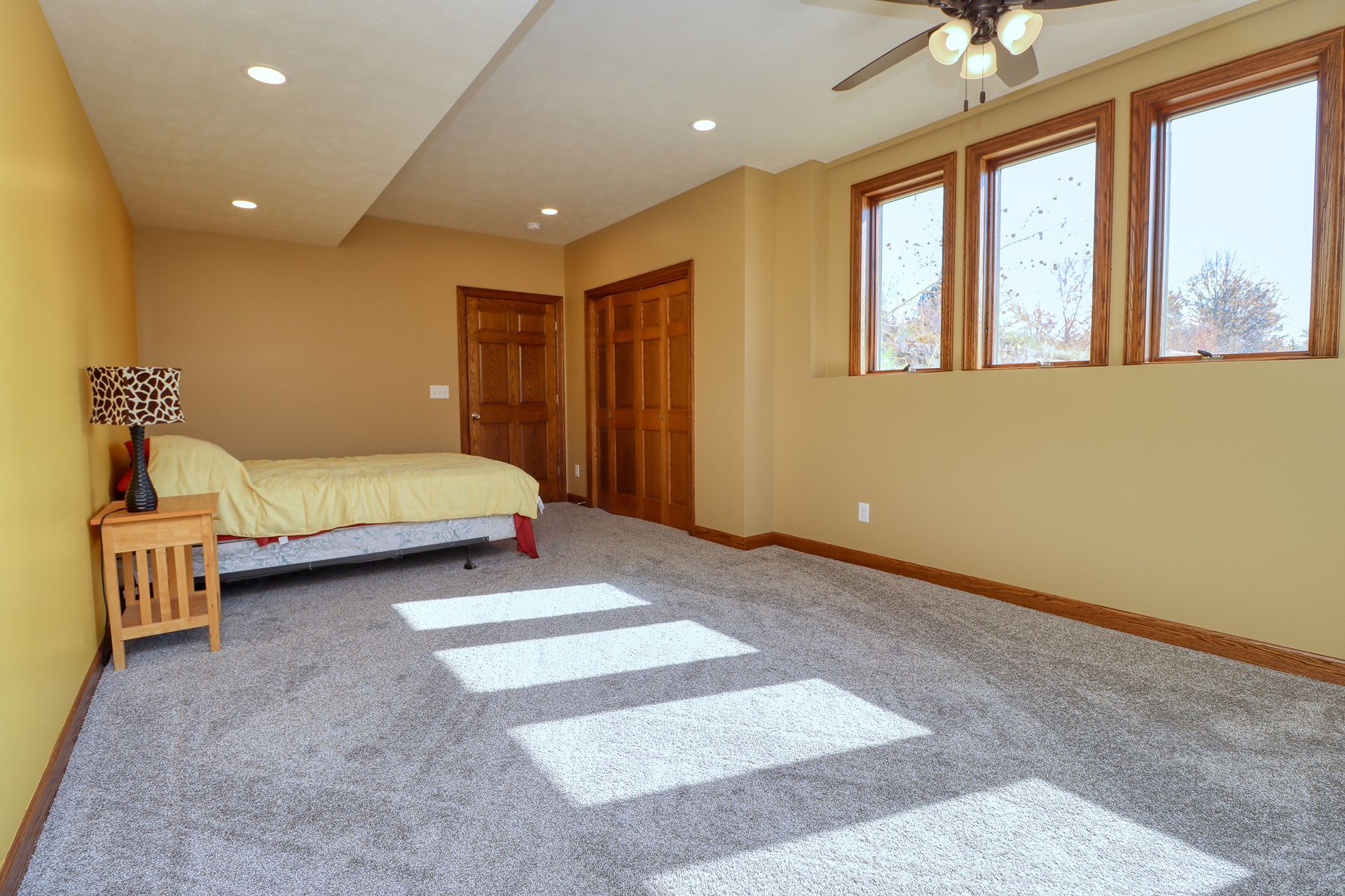 Spacious bedroom with bed, wooden furniture, large windows, and sunlight on gray carpet.