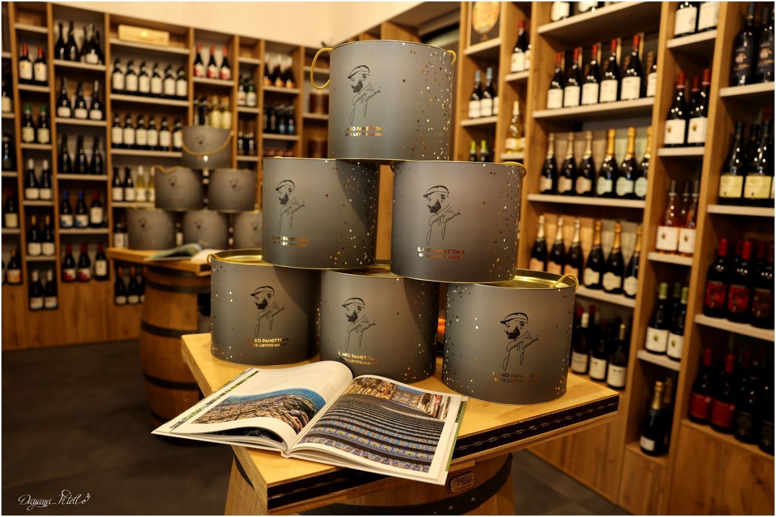 In un'enoteca, sullo sfondo di scaffali pieni di bottiglie, sono impilati dei contenitori regalo grigio scuro su una botte di legno.