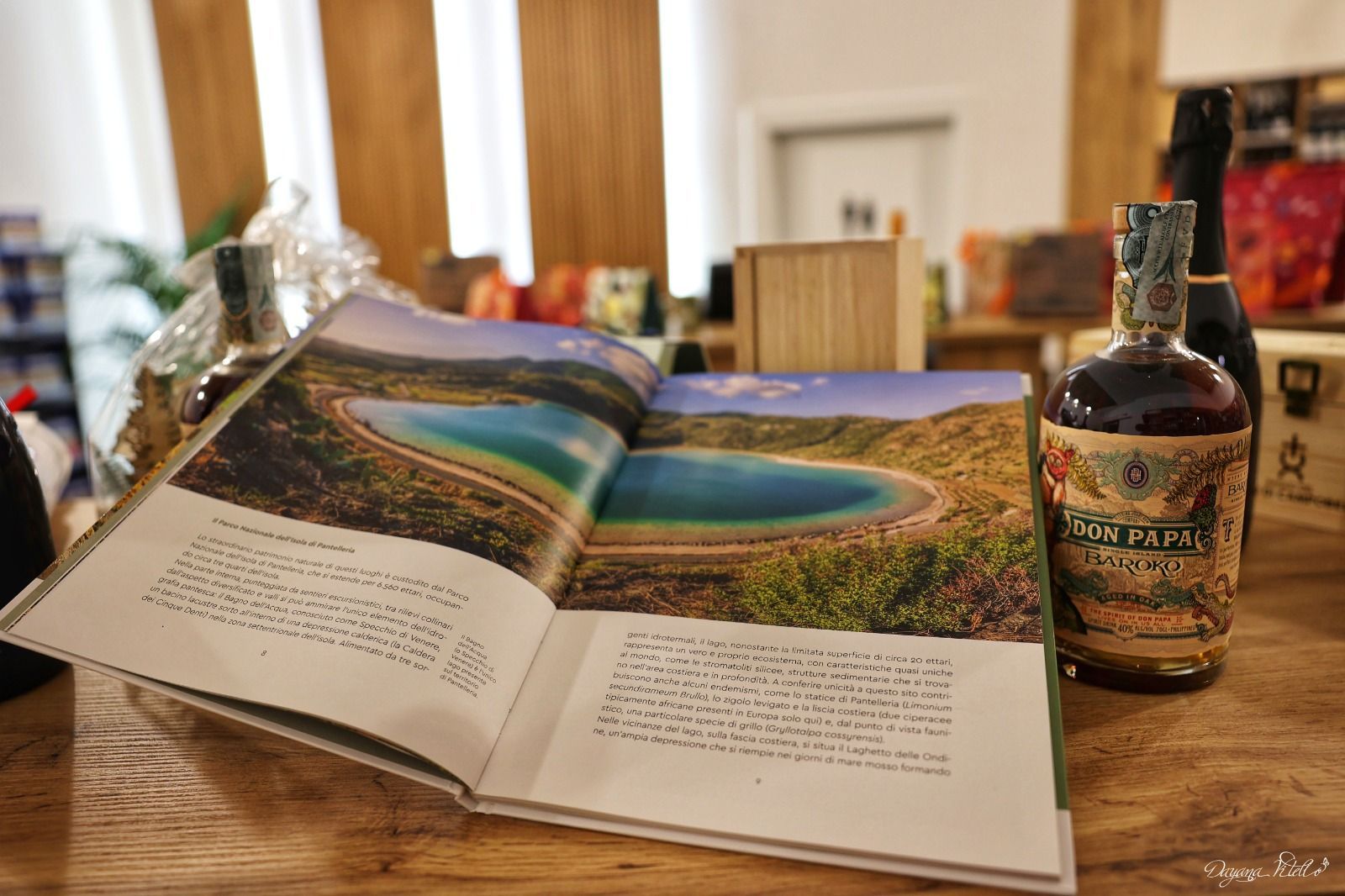 Un libro aperto, con la vivace fotografia di un lago, è appoggiato su un tavolo di legno accanto a una bottiglia di liquore scuro.