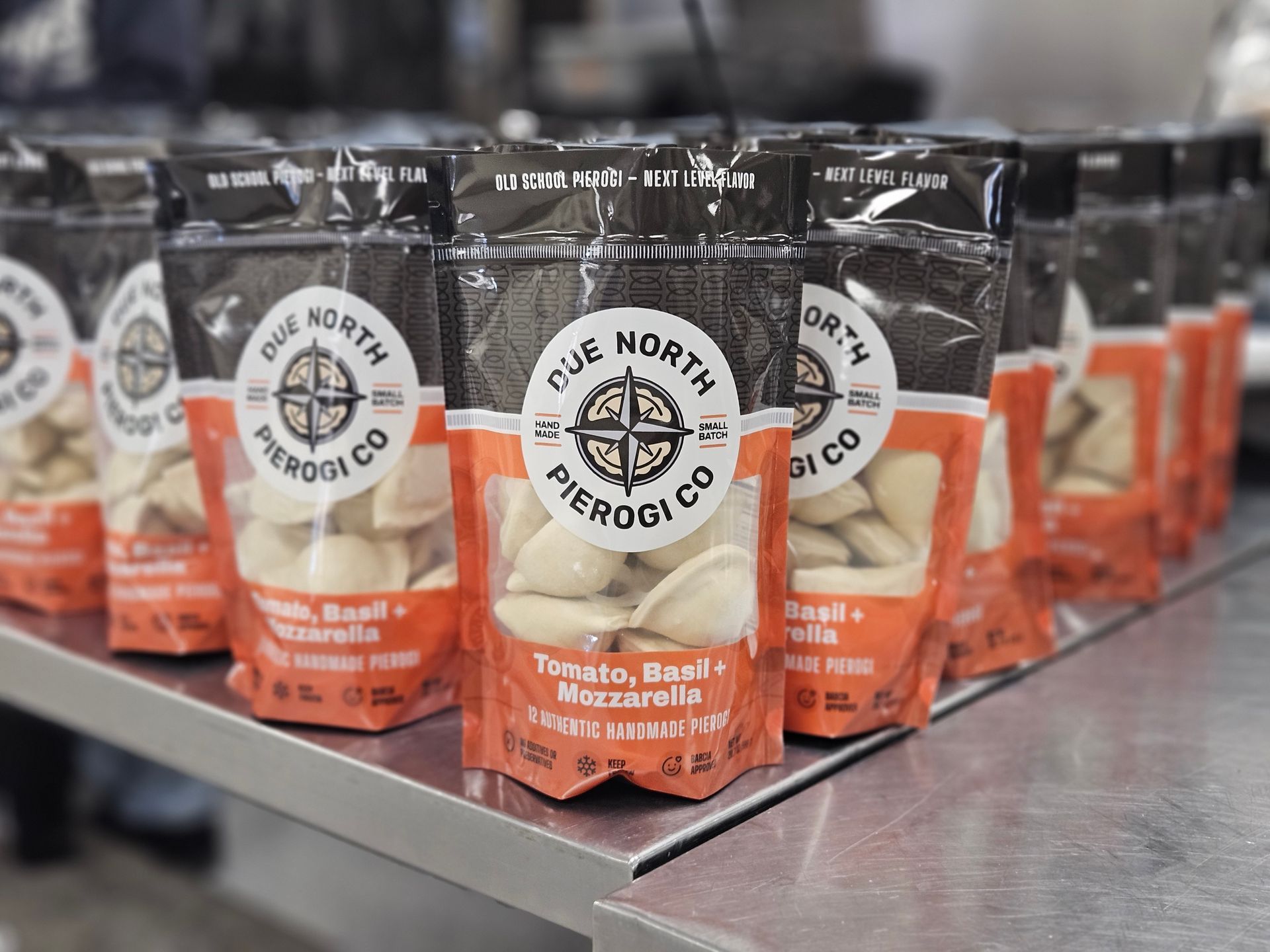Rows of Cue North pesto bags on a metal counter, with black-and-orange packaging in focus.