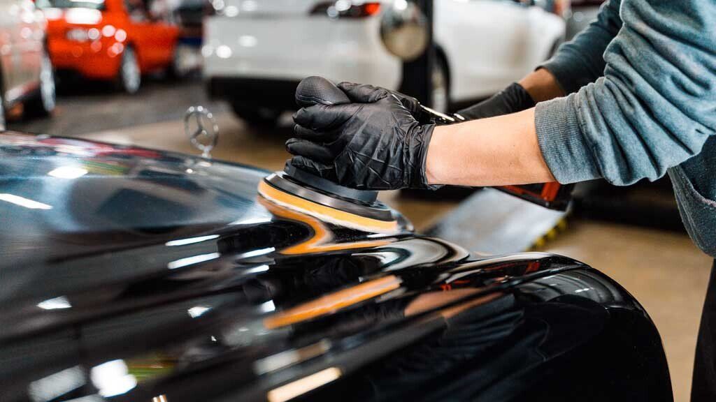 Person polishing black car with a power buffer, wearing black gloves, in a garage.