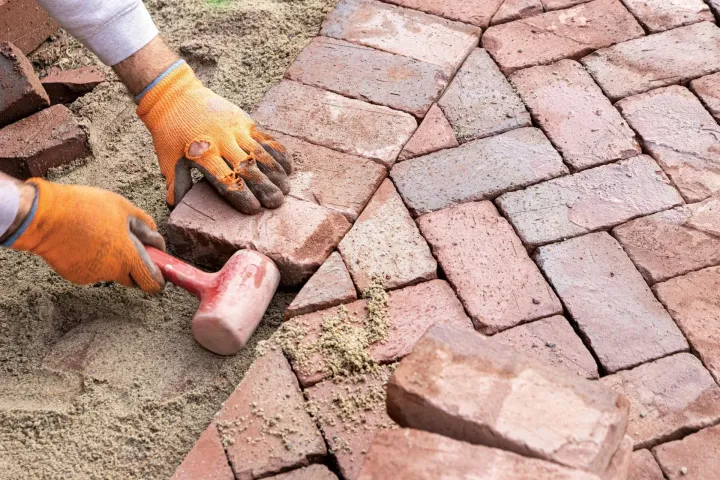 Hands laying red brick pavers with a rubber mallet on a dirt surface