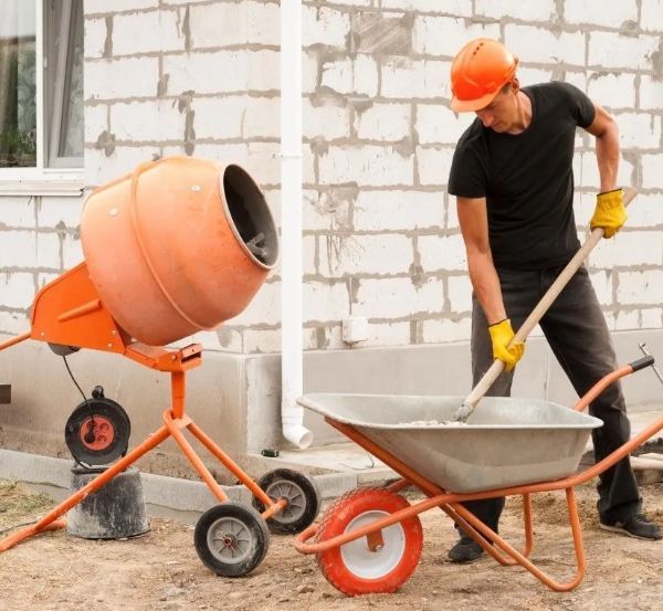 Worker in orange hard hat mixes concrete in a wheelbarrow beside a cement mixer on a construction site