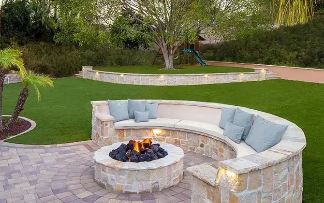 Curved stone fire pit seating area with light blue cushions in a landscaped backyard
