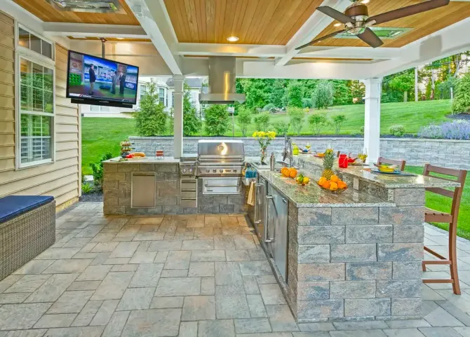Covered outdoor kitchen and bar with stone counters, TV, grill, and bar stools on a patio