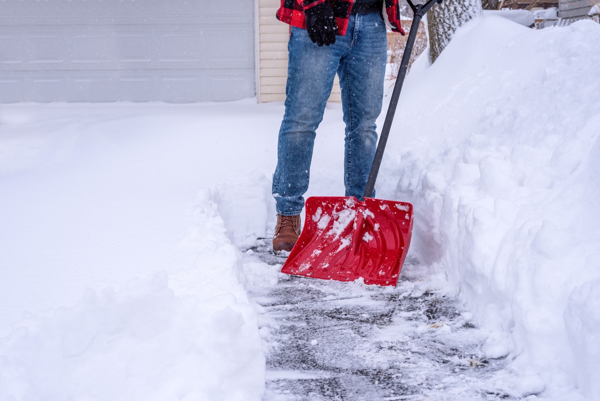 Person shoveling snow from a brick path in front of a garage, wearing a plaid shirt and jeans; winter.