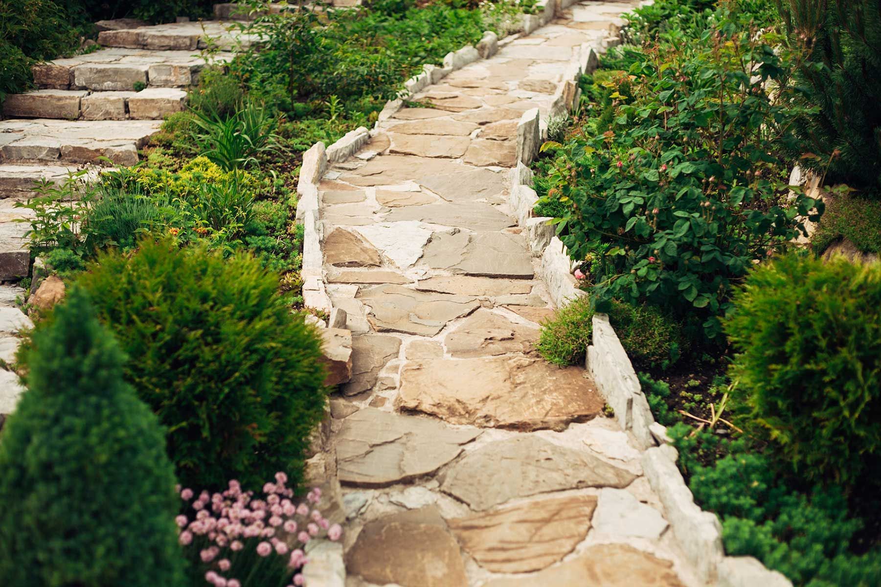 Stone path winds through a garden, flanked by lush greenery and stone stairs.