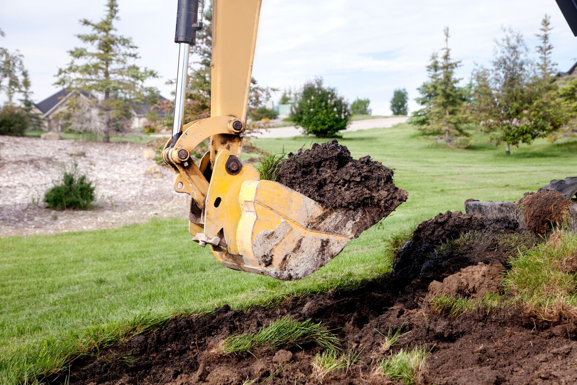 Excavator bucket filled with dark soil, digging into a grassy hillside.