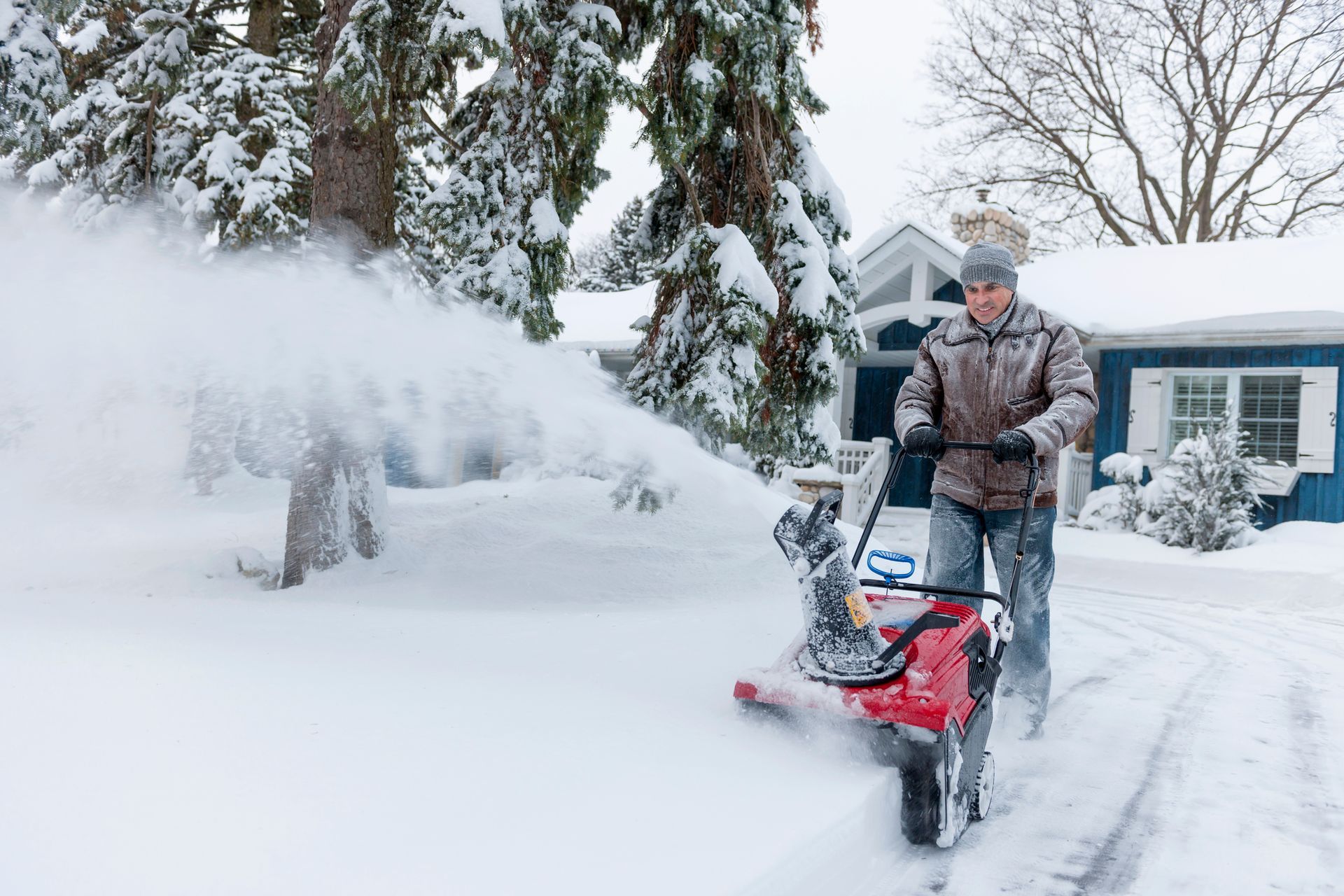 Man using a red snowblower to clear a snowy driveway near a blue house and snowy trees.