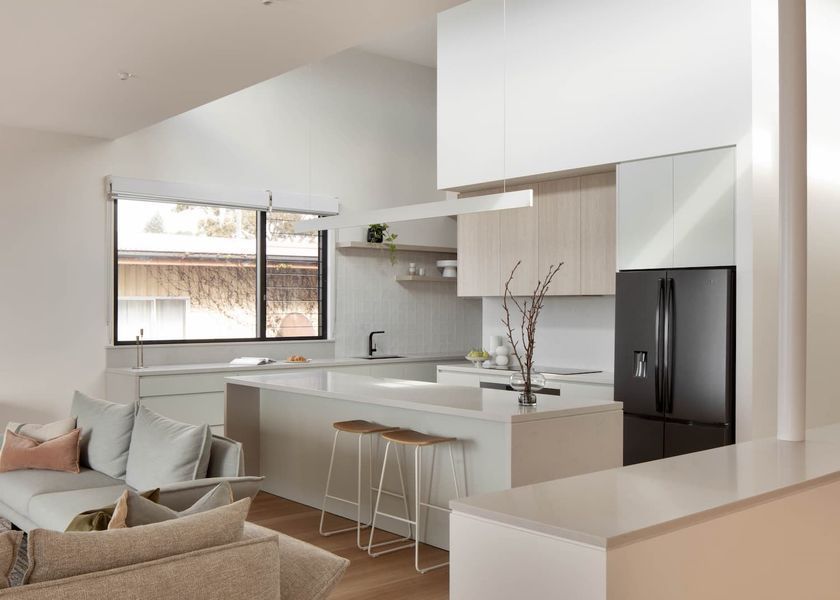 Modern kitchen with white cabinets, light wood accents, island, and black refrigerator; view includes living room sofa.