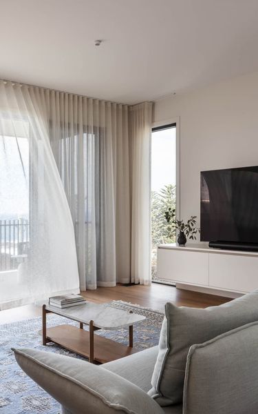 Living room with sheer curtains blowing, couch, coffee table, and TV on a white wall.