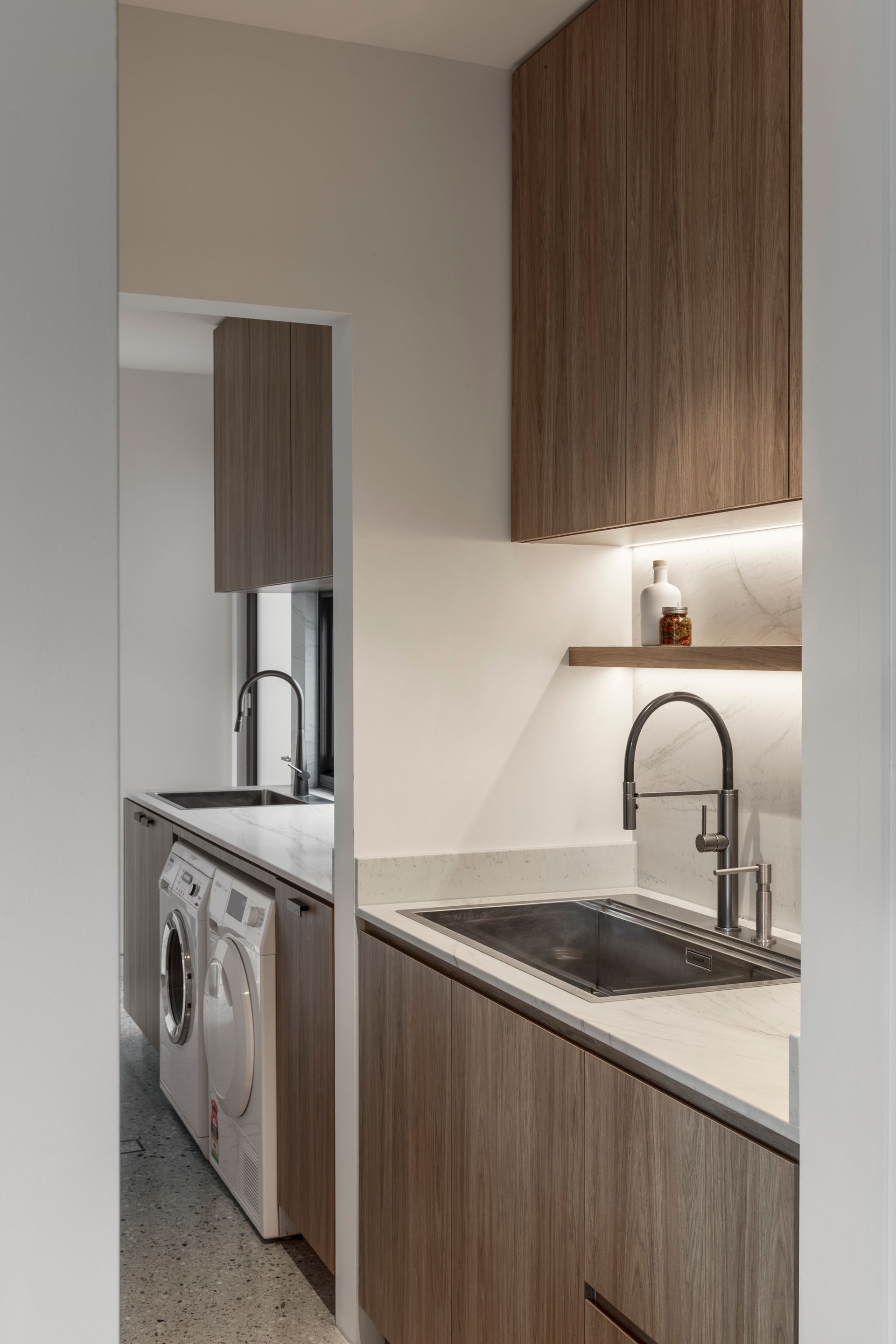 Narrow laundry room with wood cabinets, sink, and a washing machine reflected in a mirror.