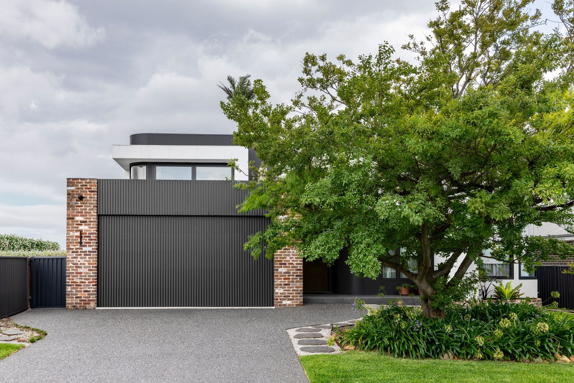 Modern house facade with dark garage door, stone pillars, gravel driveway, and a leafy tree in front