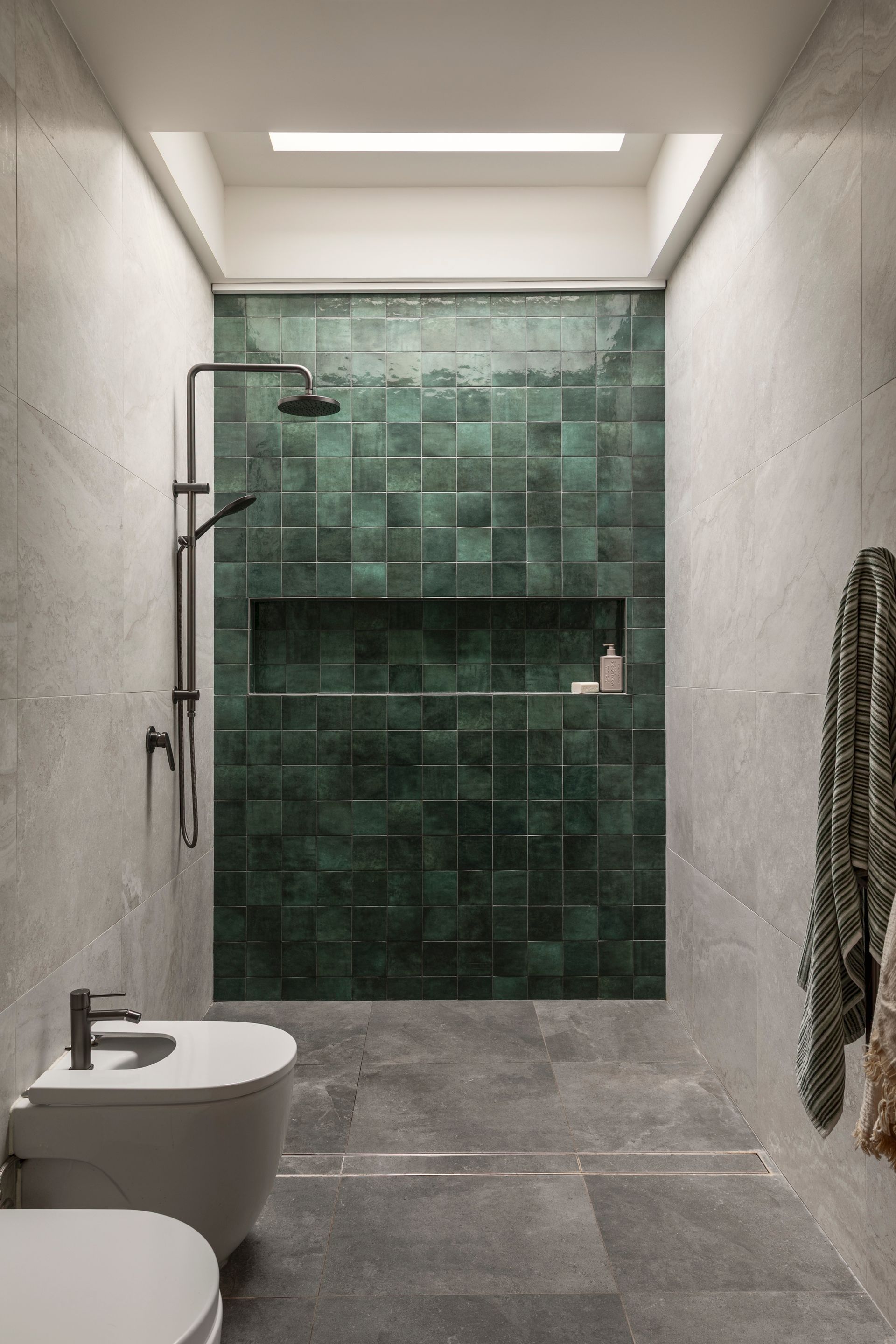 Narrow modern bathroom with gray walls, a white toilet, and a green tiled shower at the back.