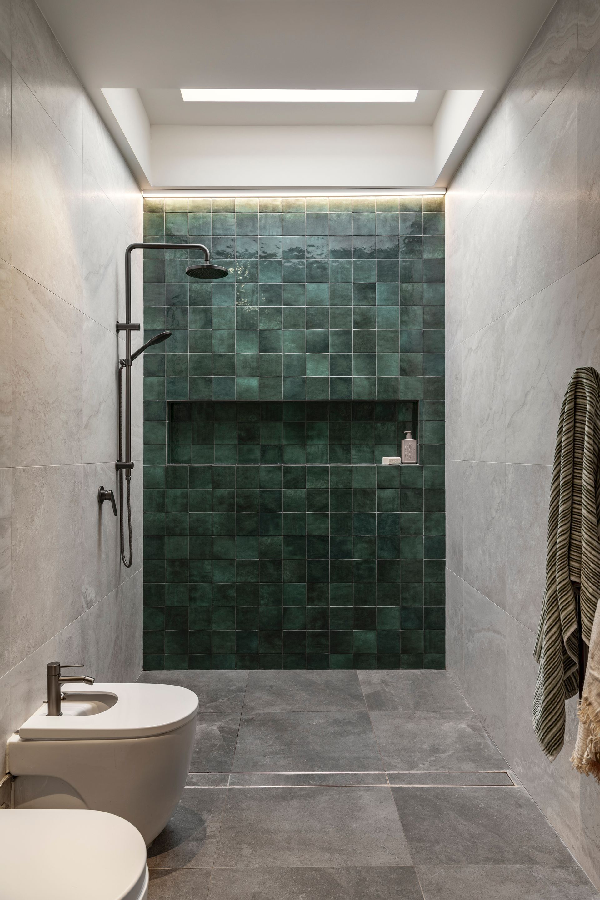 Narrow modern bathroom with gray walls, green tile shower, wall-mounted toilet, and overhead light