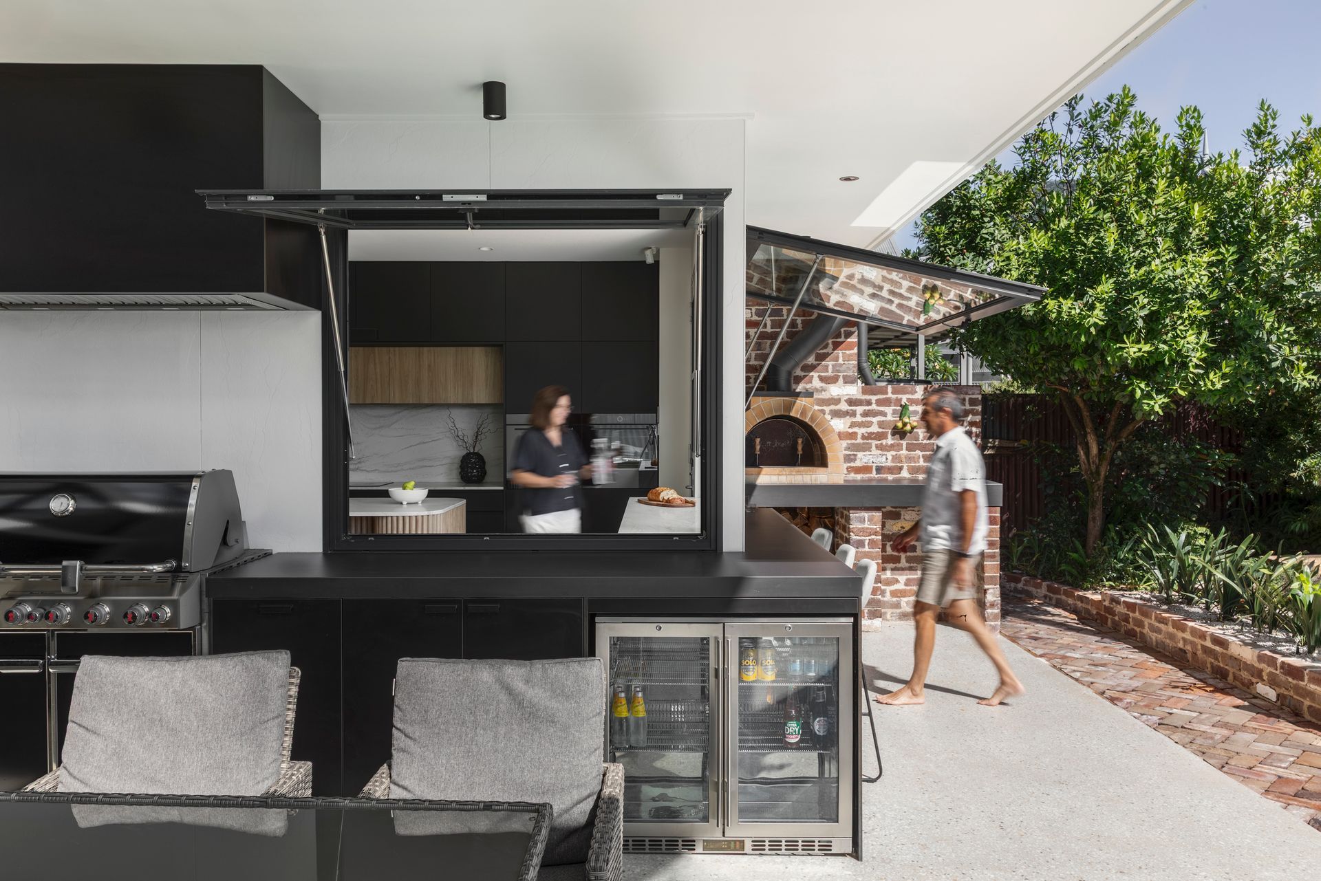 Outdoor patio bar with a person serving drinks at a modern black counter beside a grill and lounge chairs