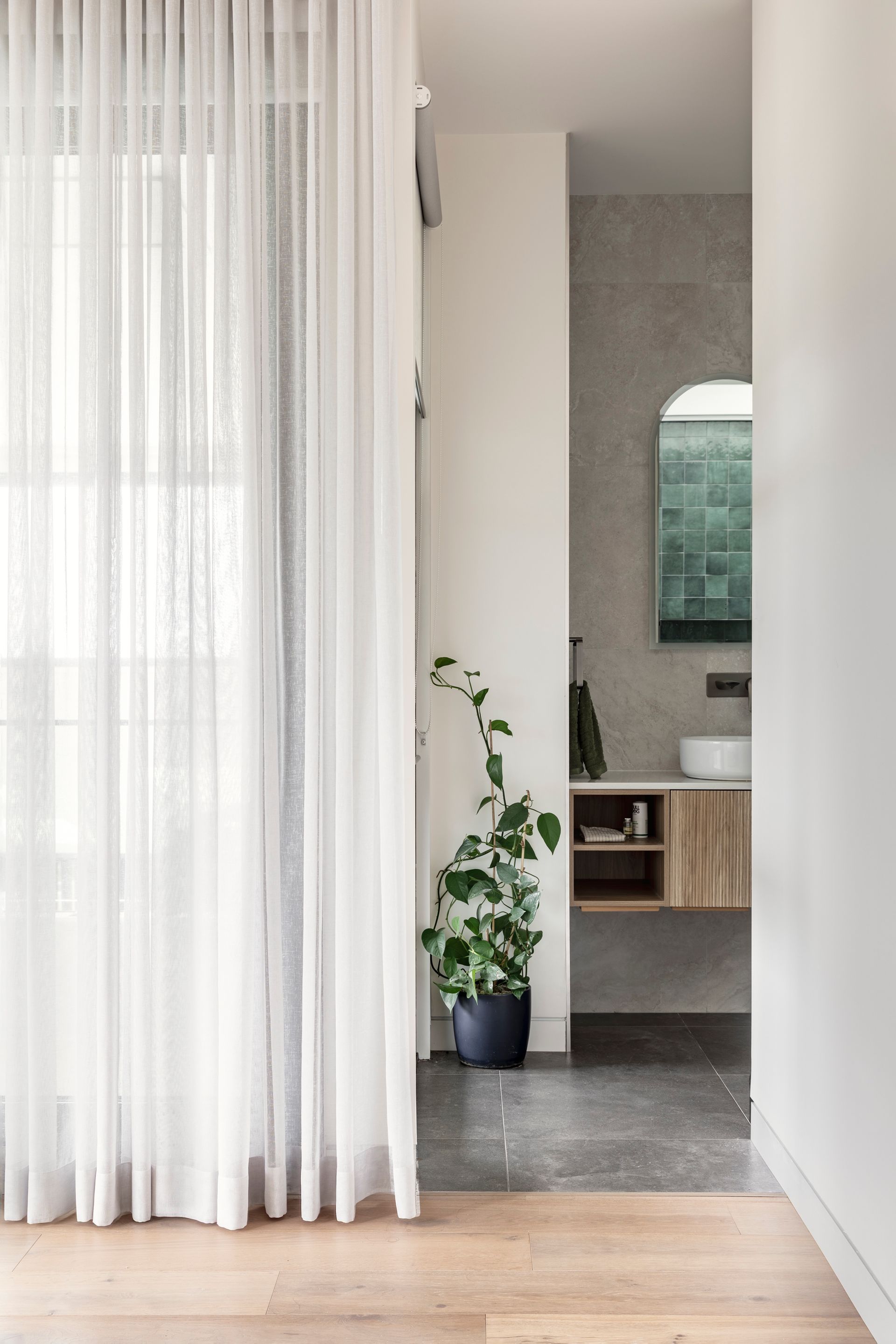Bright hallway with sheer curtains, a potted plant, and a view into a tiled bathroom vanity area.