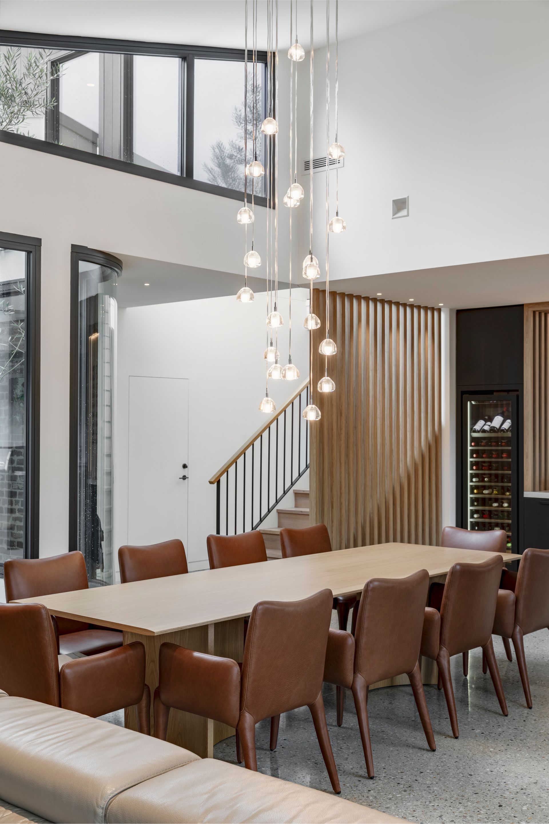 Modern dining room with long wooden table, brown chairs, and hanging pendant lights under a high ceiling
