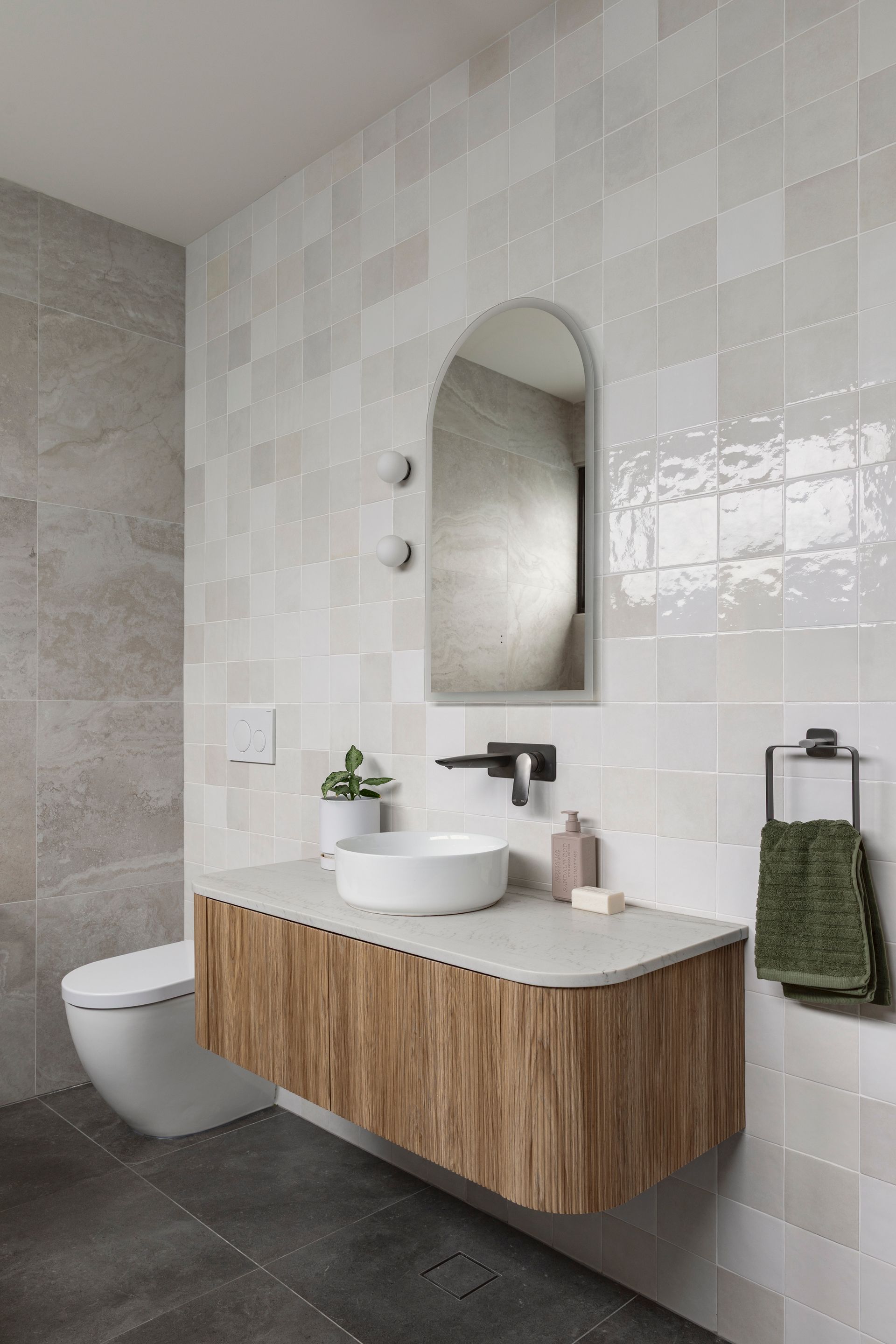 Modern bathroom with a wood vanity, round sink, oval mirror, toilet, and tiled walls