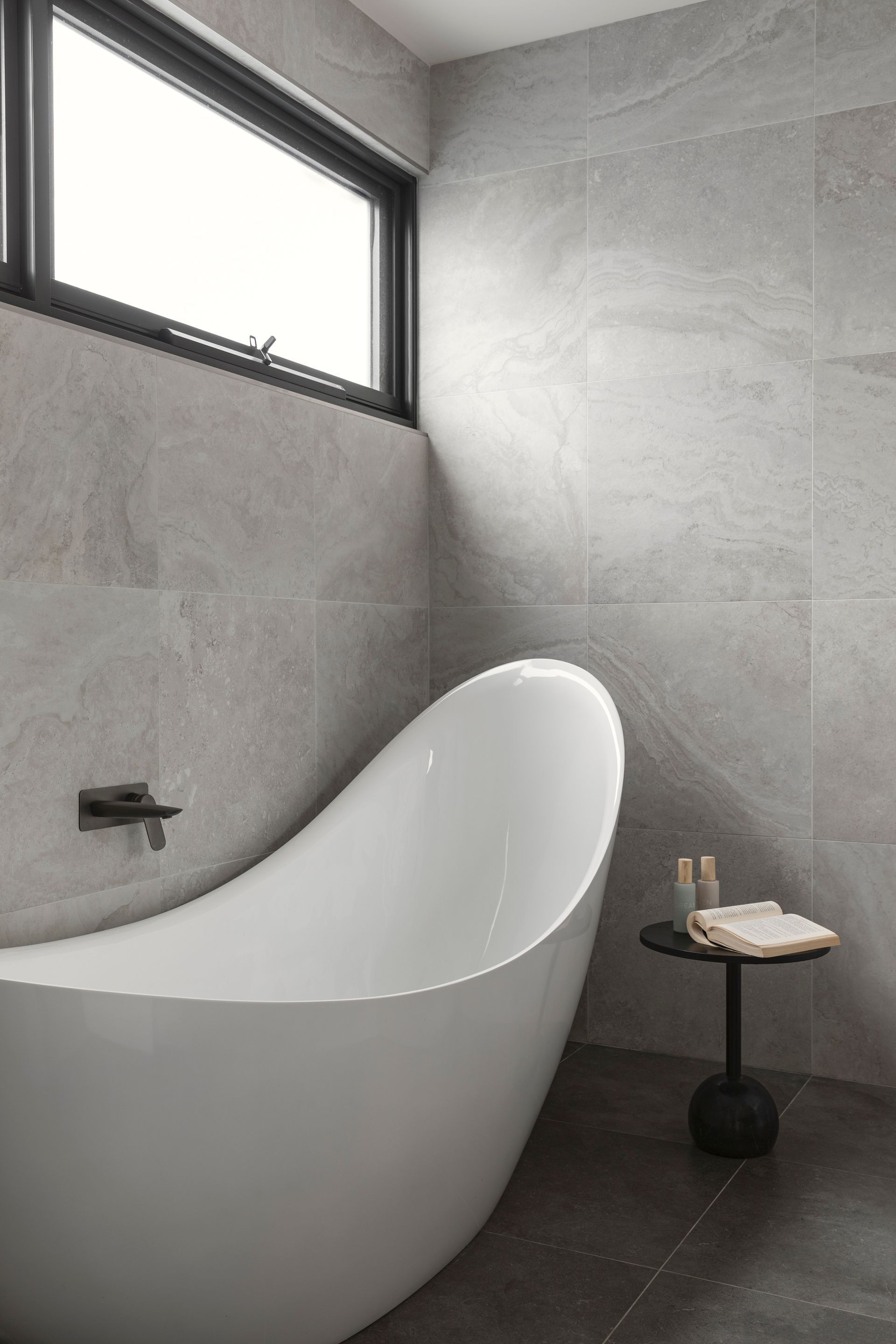 Modern bathroom with a white freestanding tub, gray tiled walls, and a small side table by the window