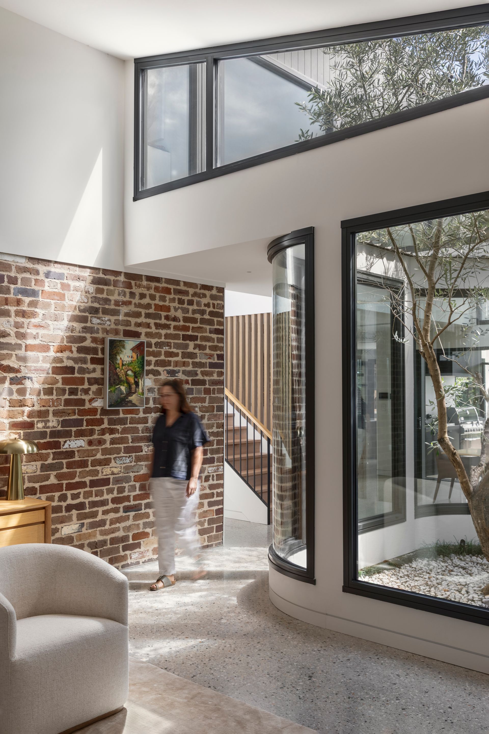 Bright modern living room with brick wall, large windows, and a person standing near the stairs
