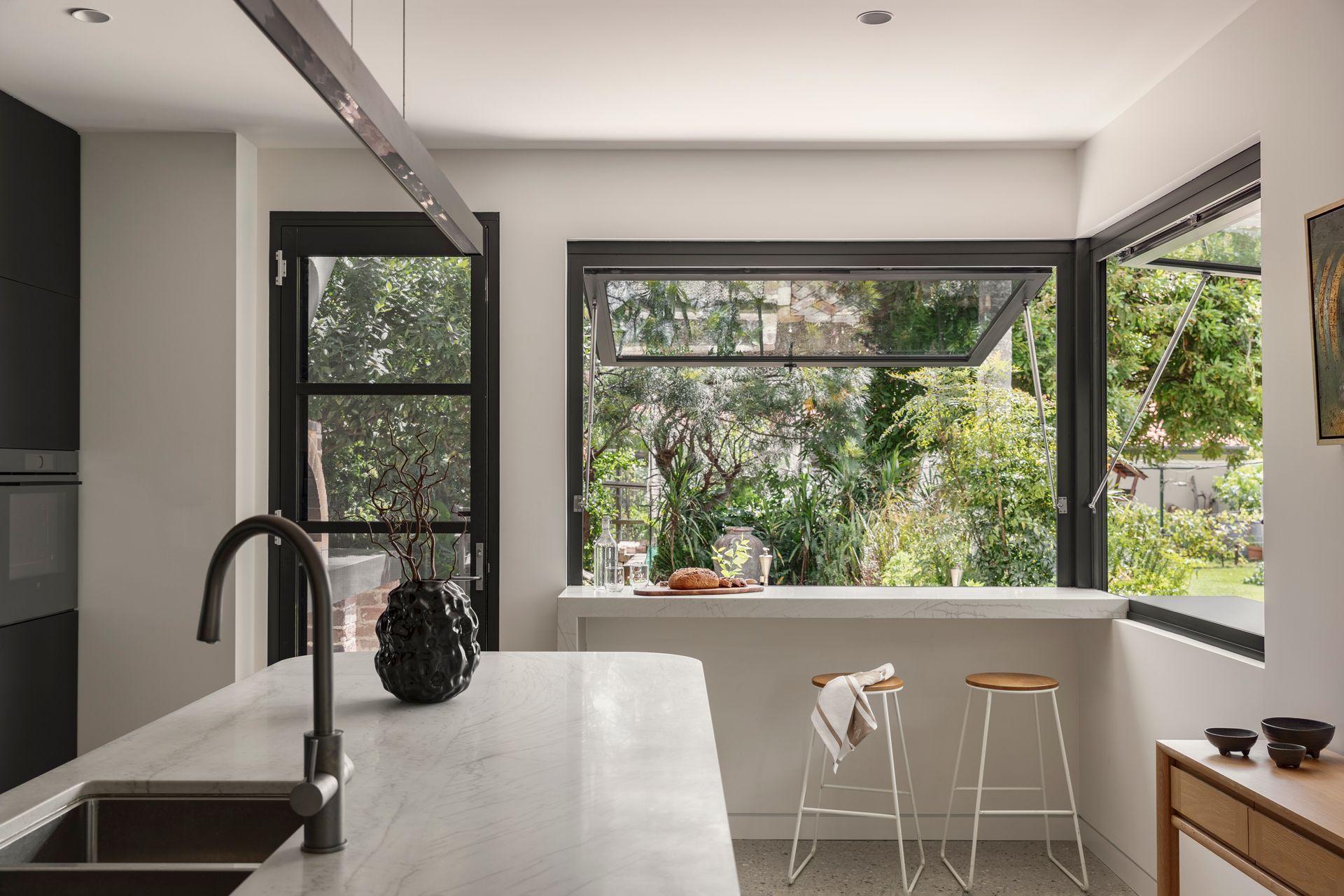 Modern kitchen with white island, black fixtures, and large windows overlooking a green garden.