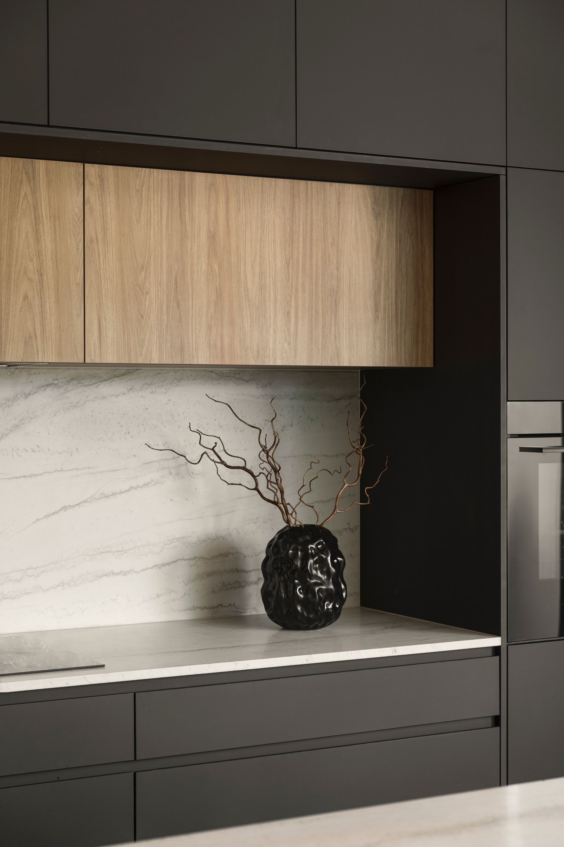 Modern kitchen nook with dark cabinets, wood shelf, marble backsplash, and a black vase with branches.