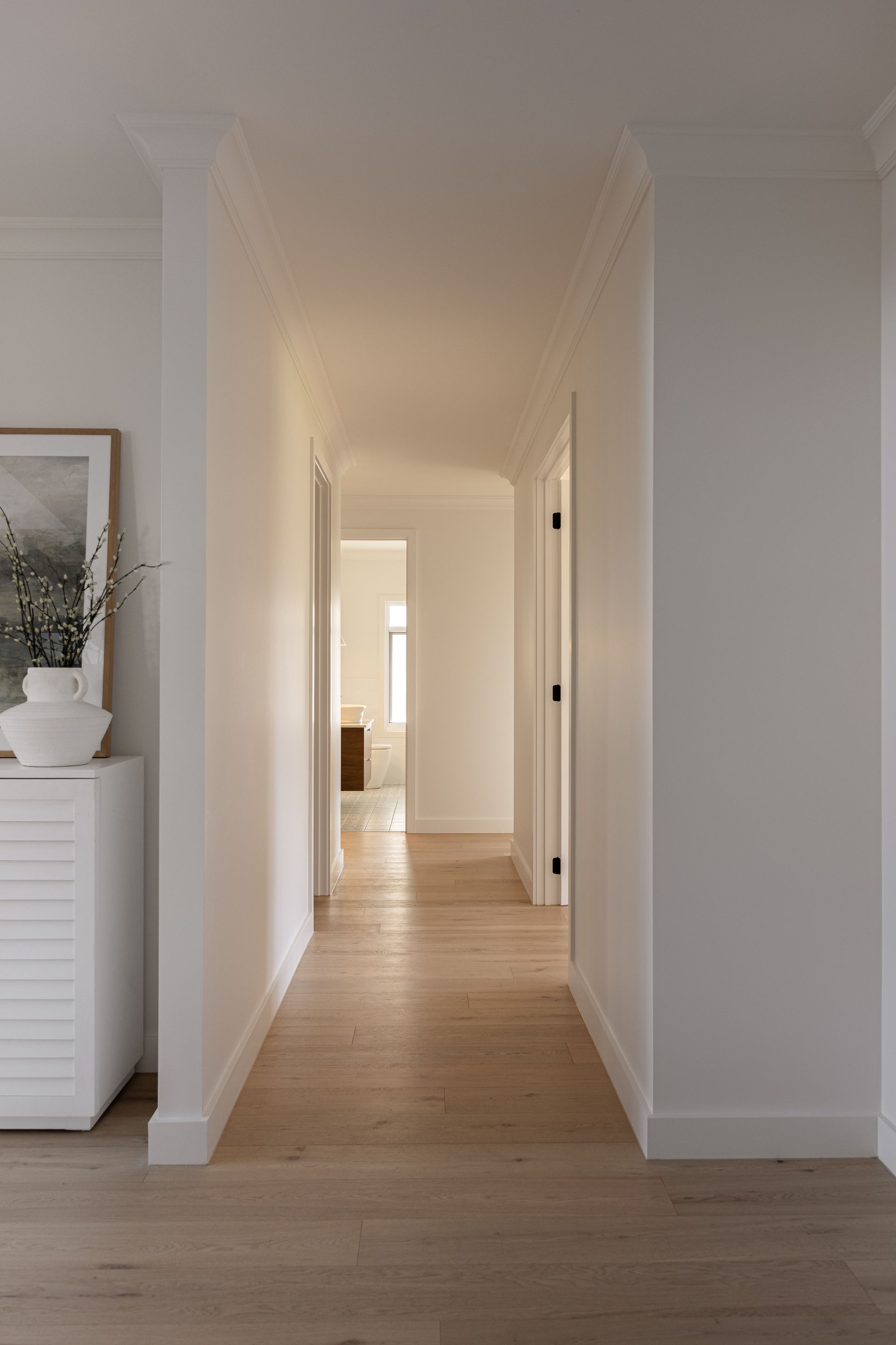 Long hallway with light wood floors, white walls, and closed doors; sunlight at the end.