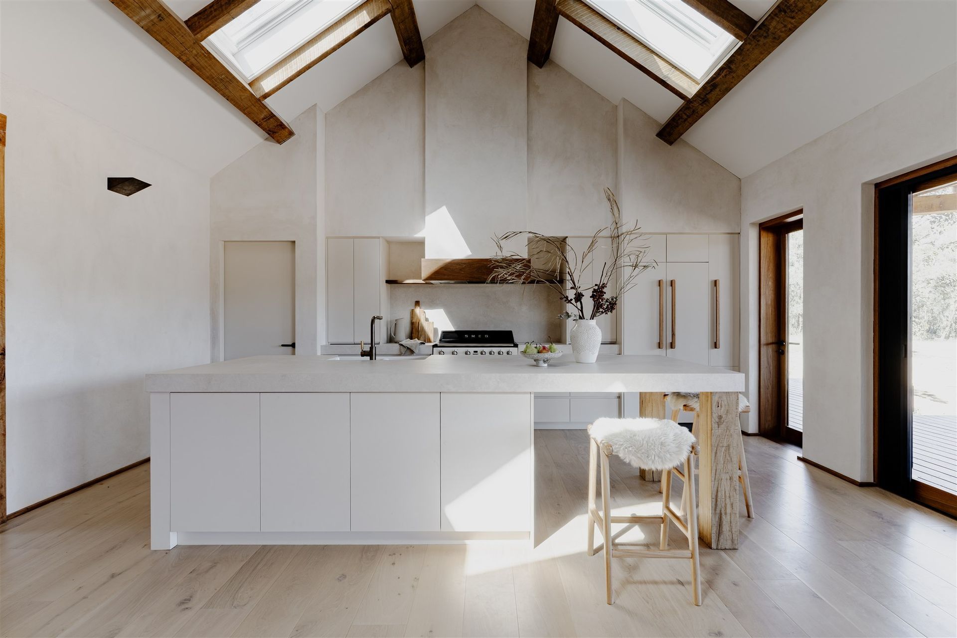 A modern, minimalist kitchen with a large white island, wooden ceiling beams, skylights, and light-toned wood flooring.