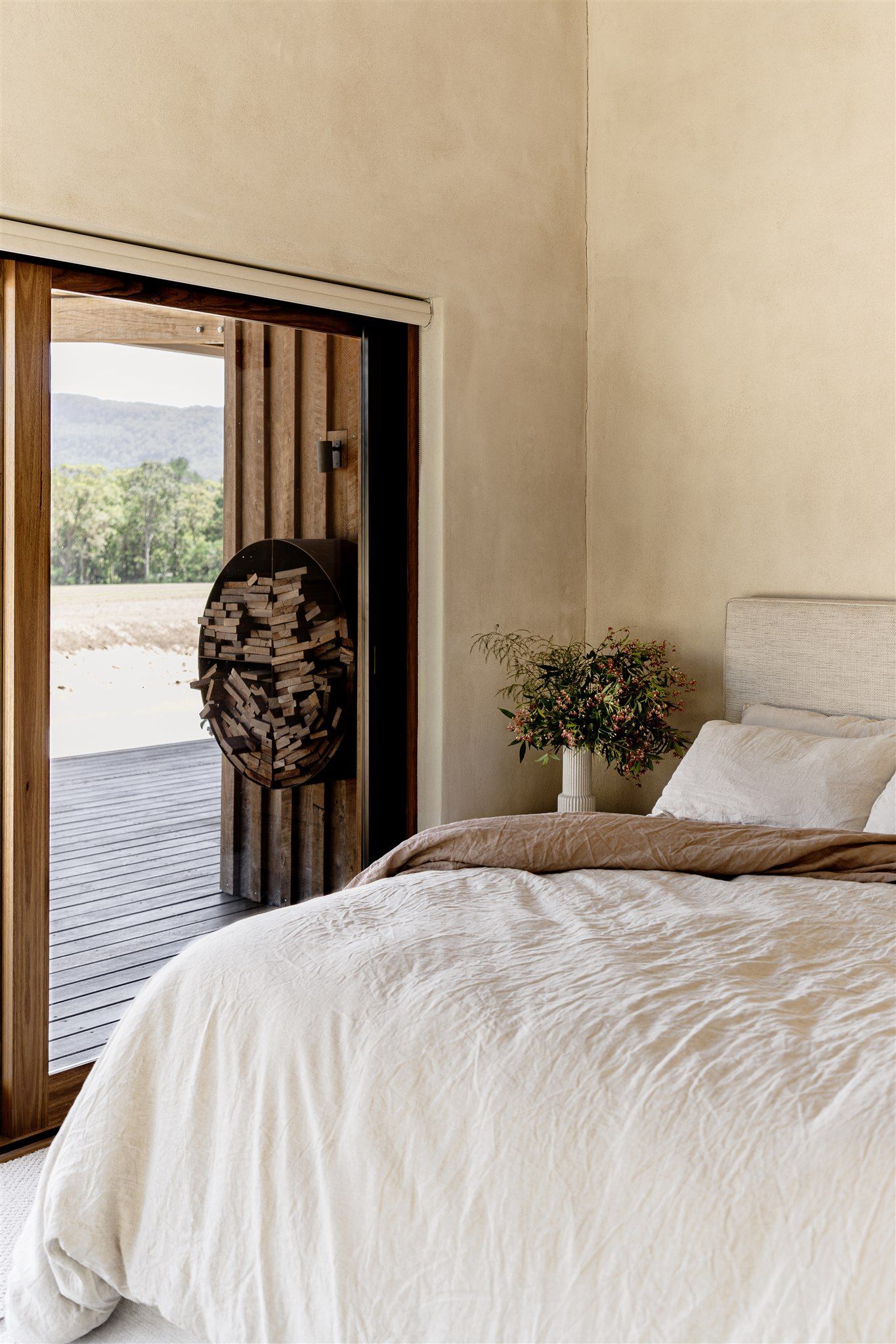 A bedroom with a textured beige wall, a bed covered in cream bedding, and an open door revealing a view of hills.