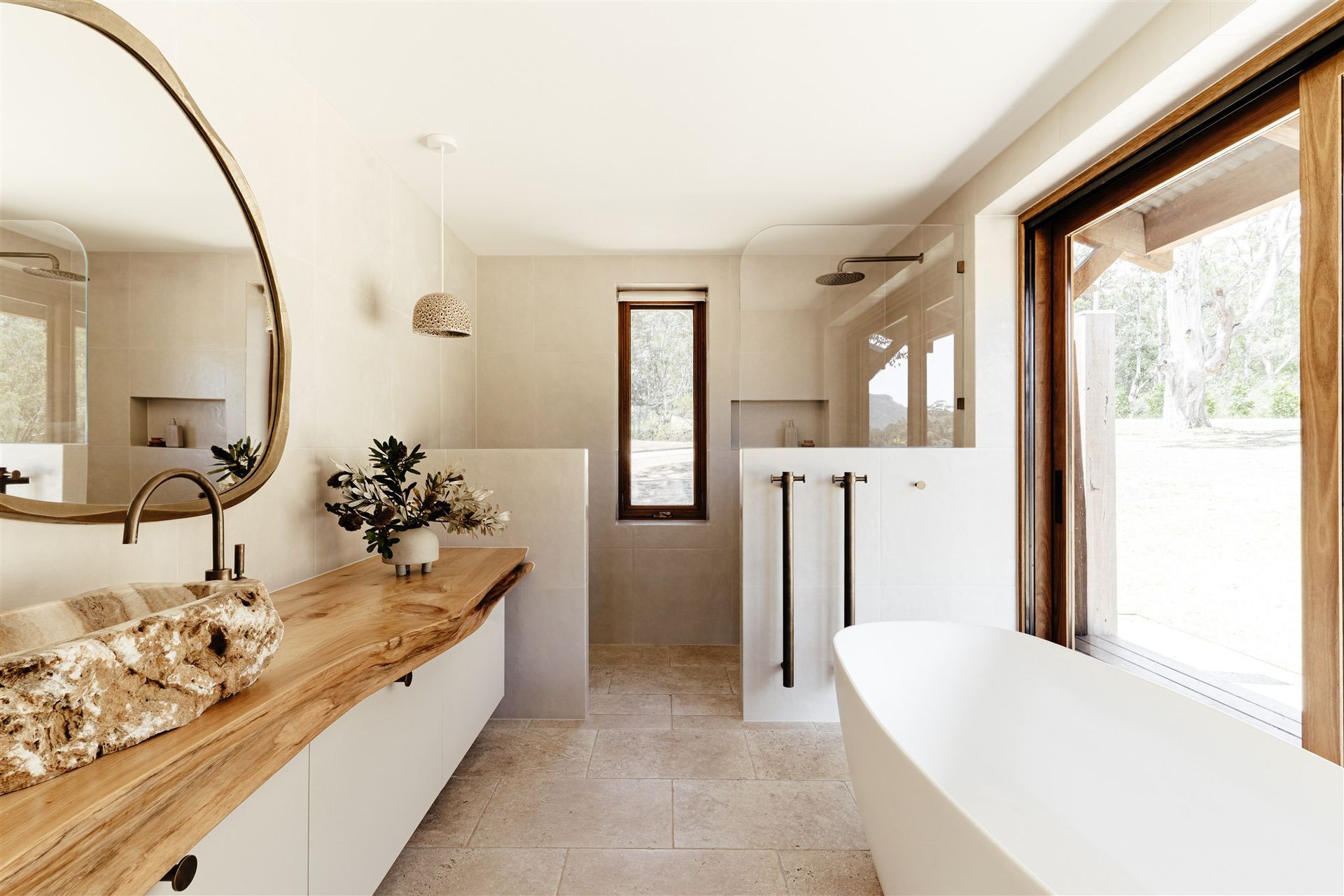 Modern bathroom with a wooden slab vanity, stone sink, oval mirror, white freestanding tub, and a large view of nature.