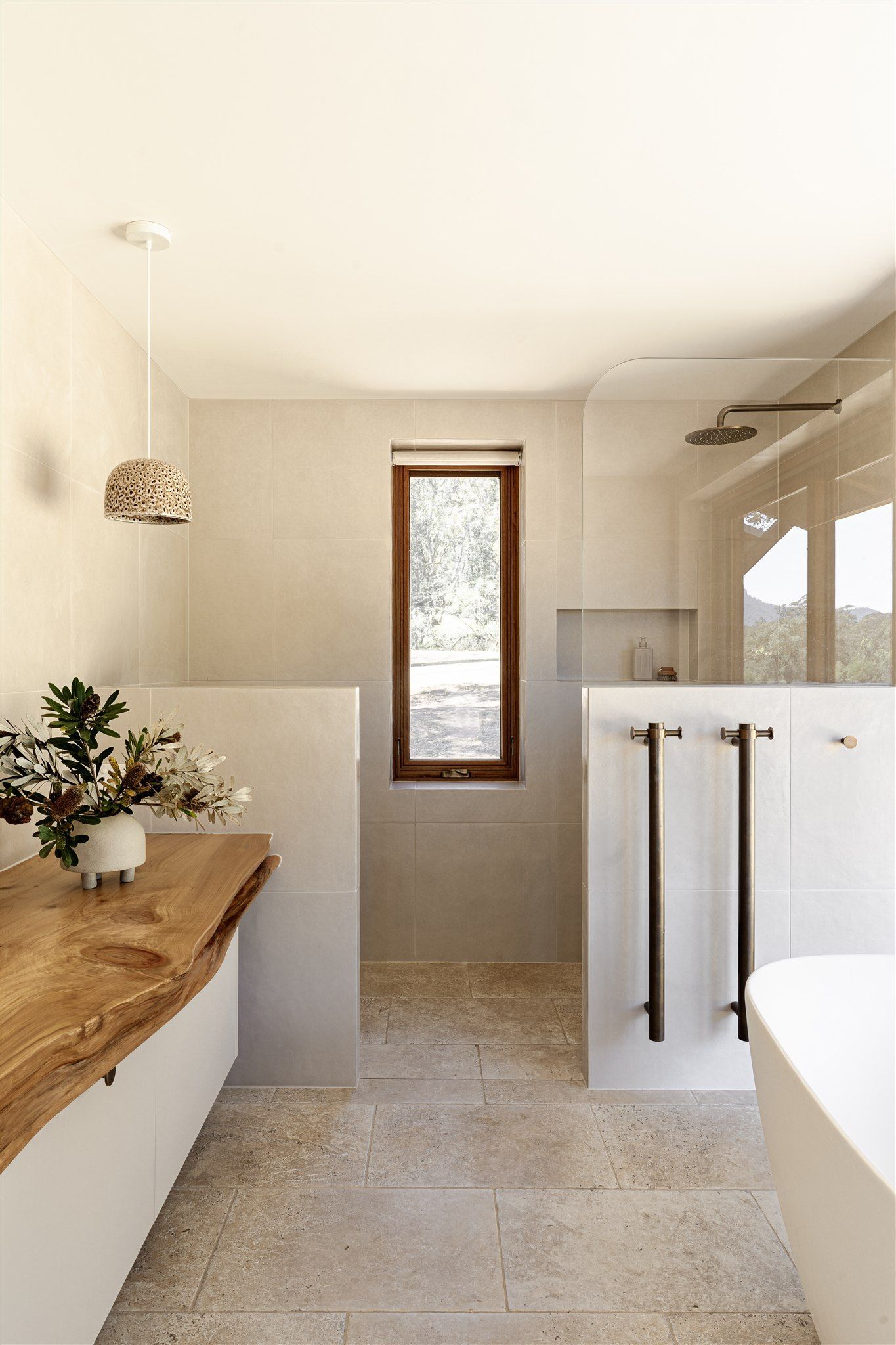 A modern bathroom with a wooden live-edge vanity, textured stone floor, freestanding tub, and a window between shower walls.