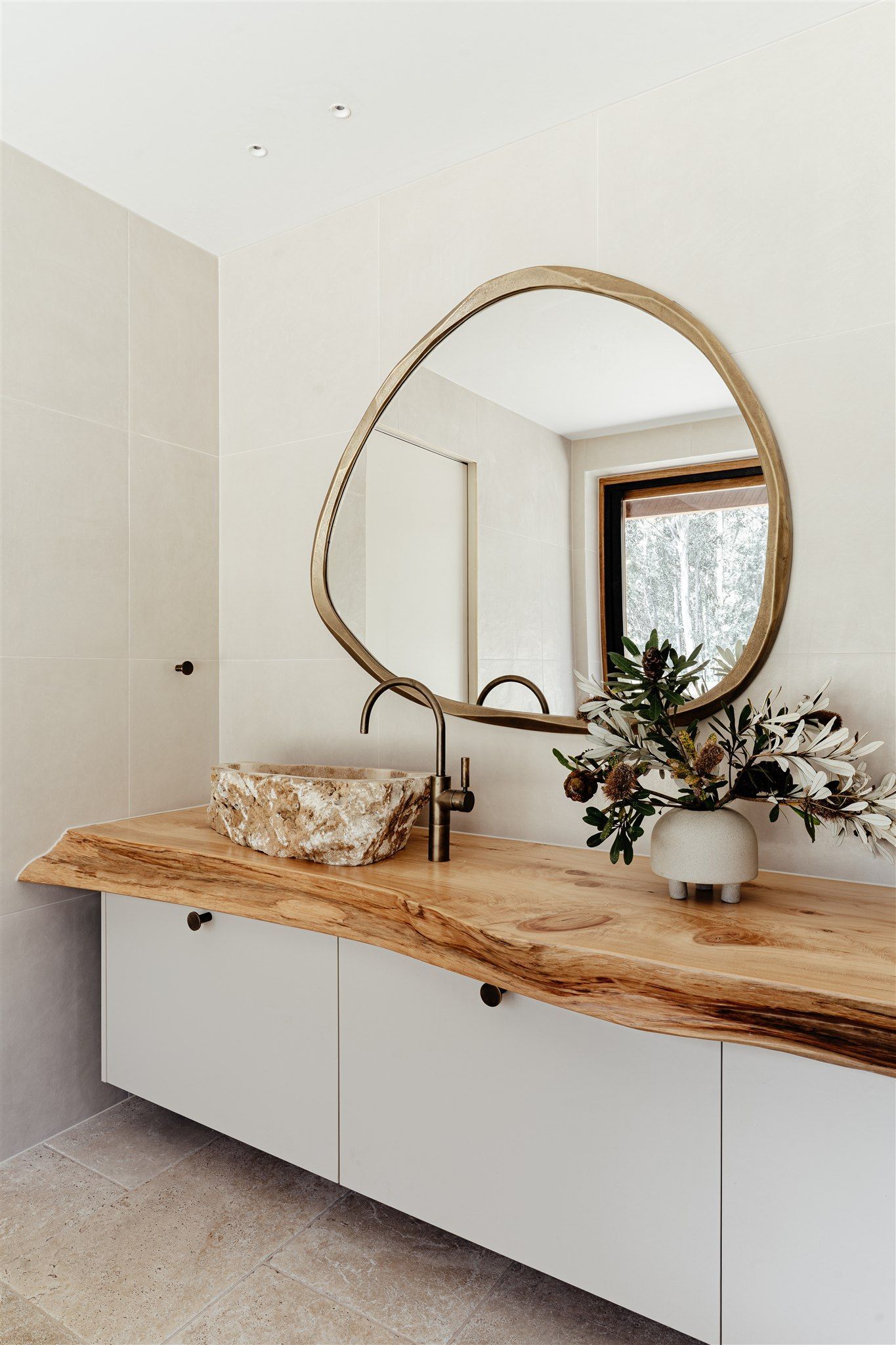 Modern bathroom vanity with a live-edge wooden countertop, organic-shaped mirror, stone vessel sink, and vase of flowers.