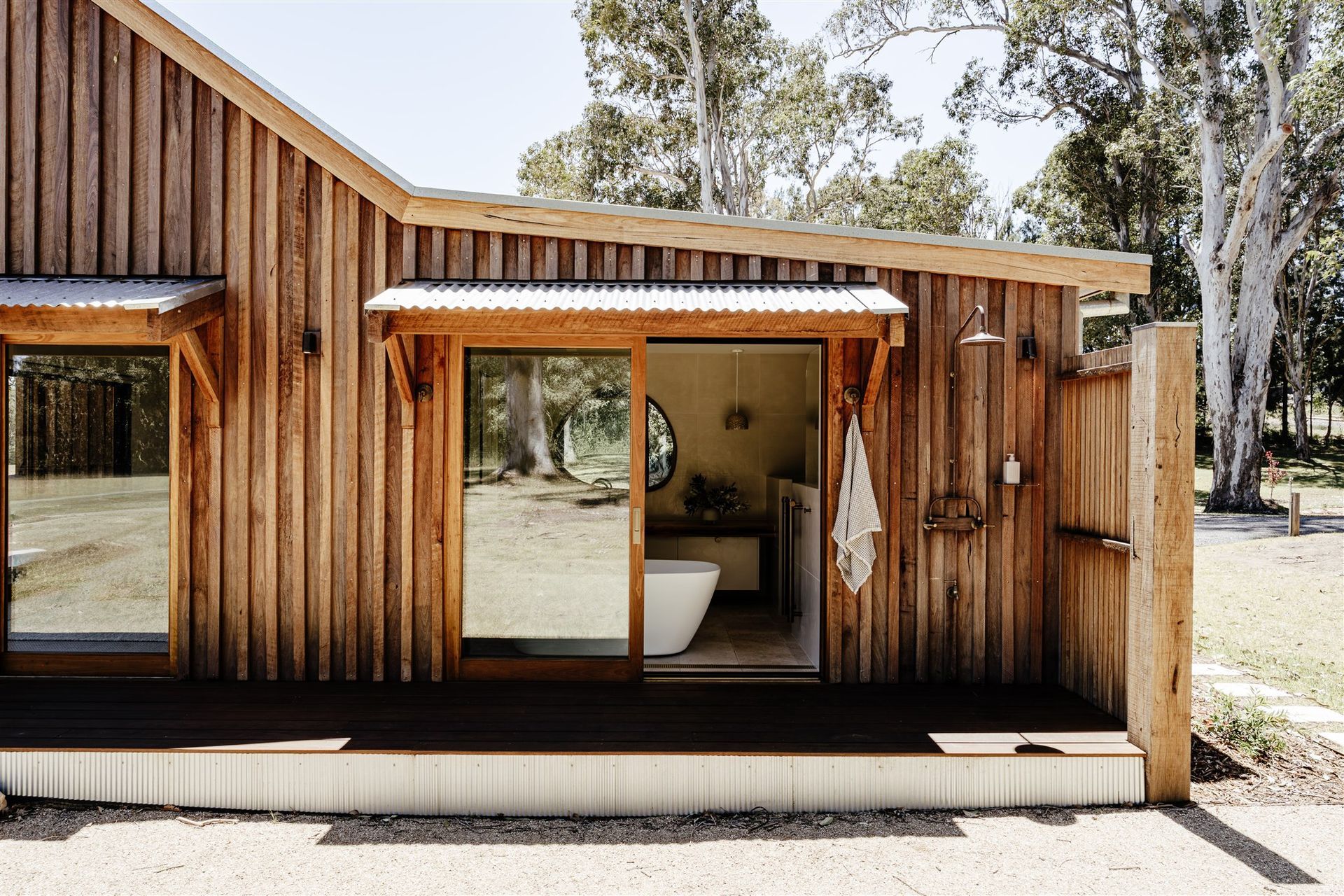 A rustic wooden cabin exterior with a glass door opening to a bathroom featuring a white tub, set in a sunny outdoor area.