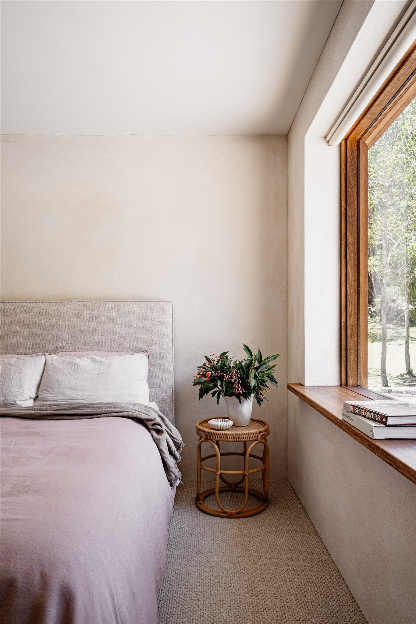 A cozy bedroom with a textured headboard, a dusty pink blanket, and a small rattan table with a plant next to a window.